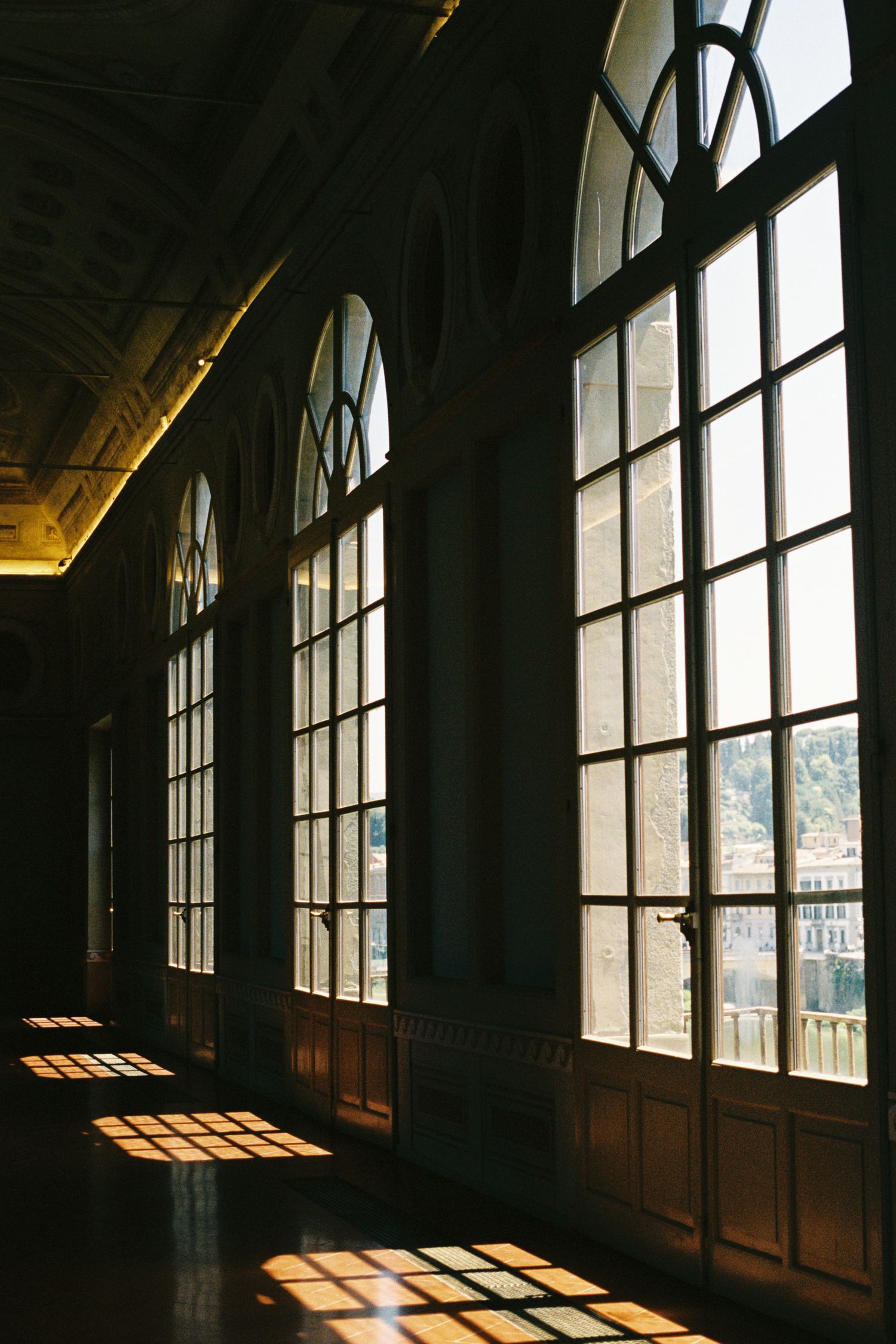 Sunlight streams through tall arched windows casting geometric shadows on the floor of an elegant historic corridor.