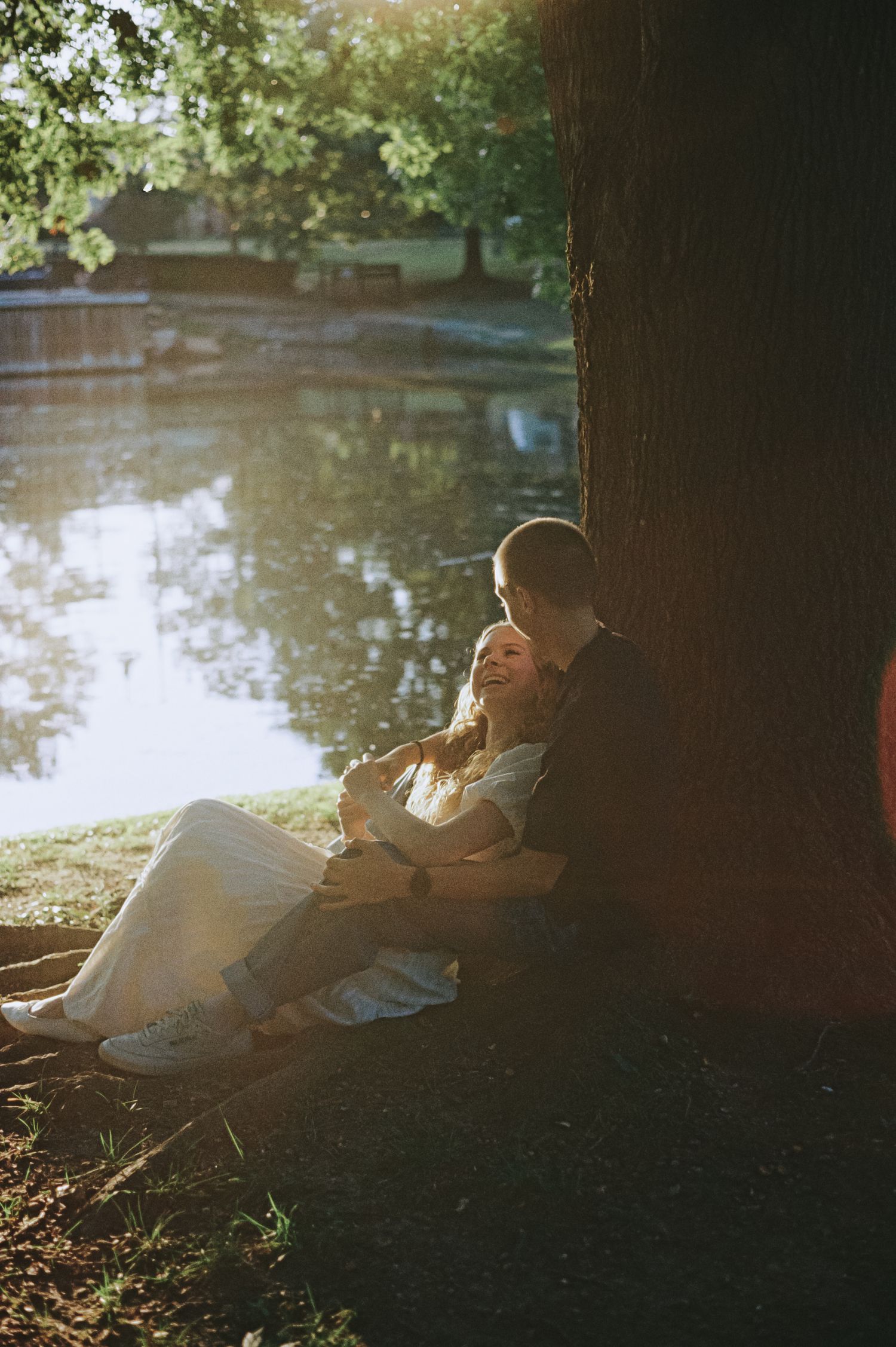 Couple sitting together leaning against a tree by a sunlit lake in a park, sharing a tender romantic moment.