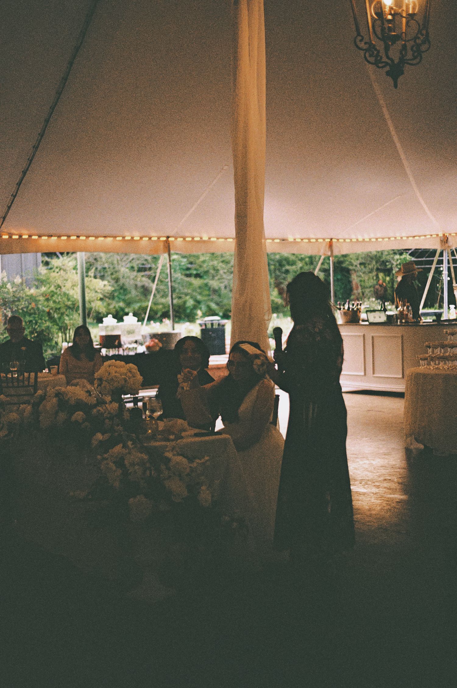 Silhouetted wedding guests gathered under an elegant tent with string lights and floral arrangements at a reception.