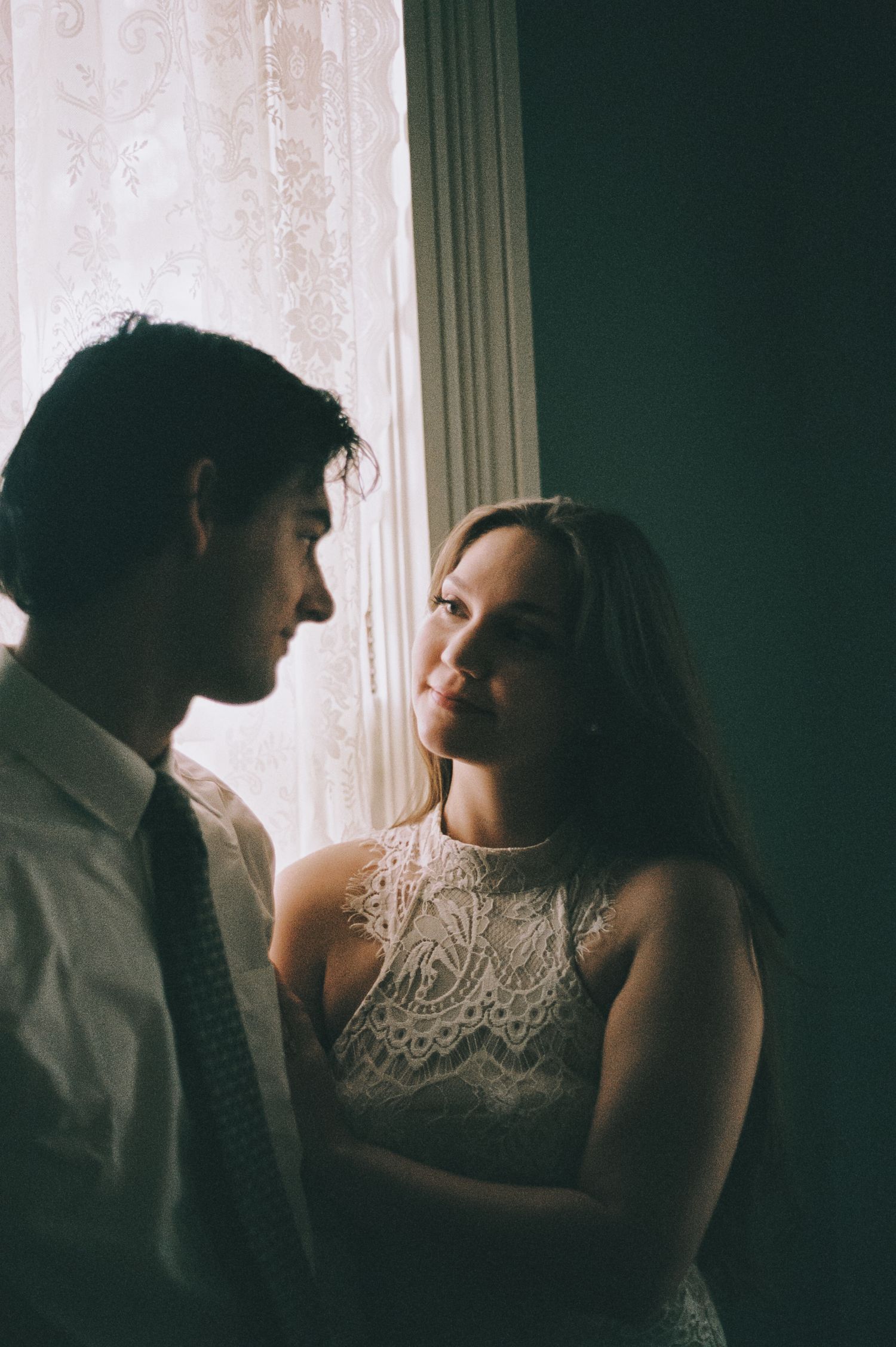 Bride in lace dress smiles at groom in tie while standing together by a window.