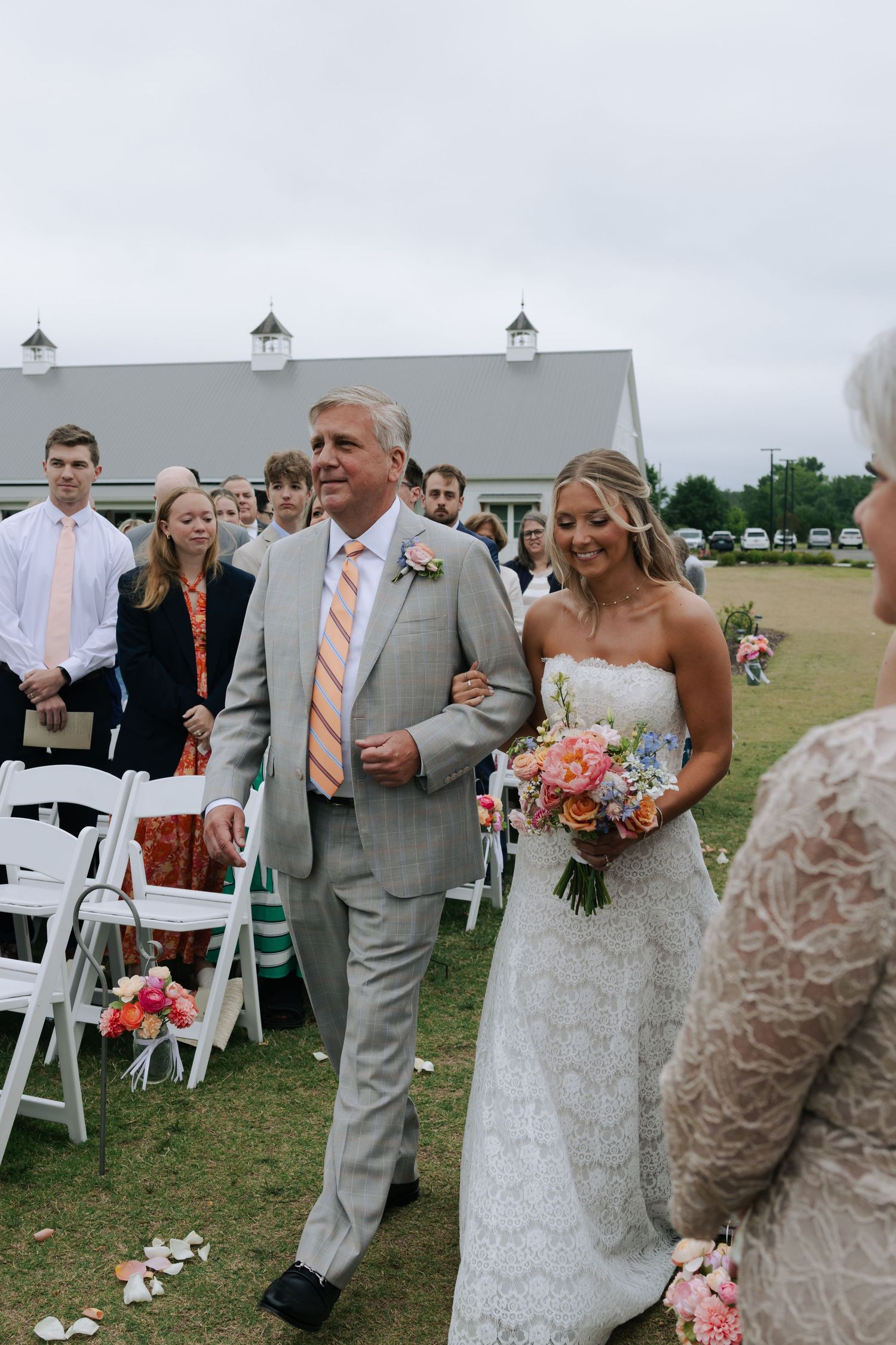 Father walks bride down outdoor wedding aisle in lace gown holding colorful bouquet.