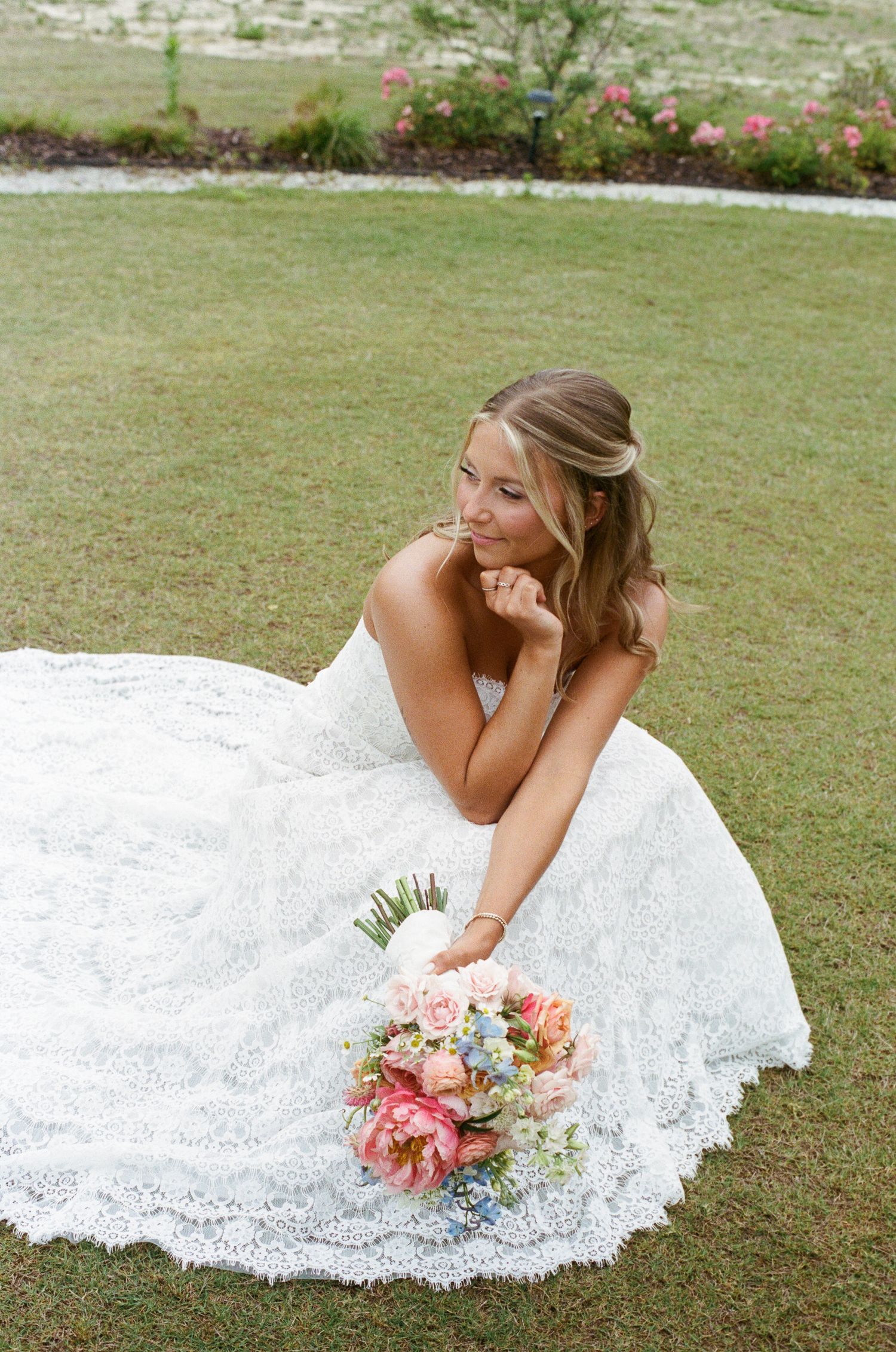 Bride in a flowing white lace gown sitting on grass smiling with a colorful pink and blue bridal bouquet.