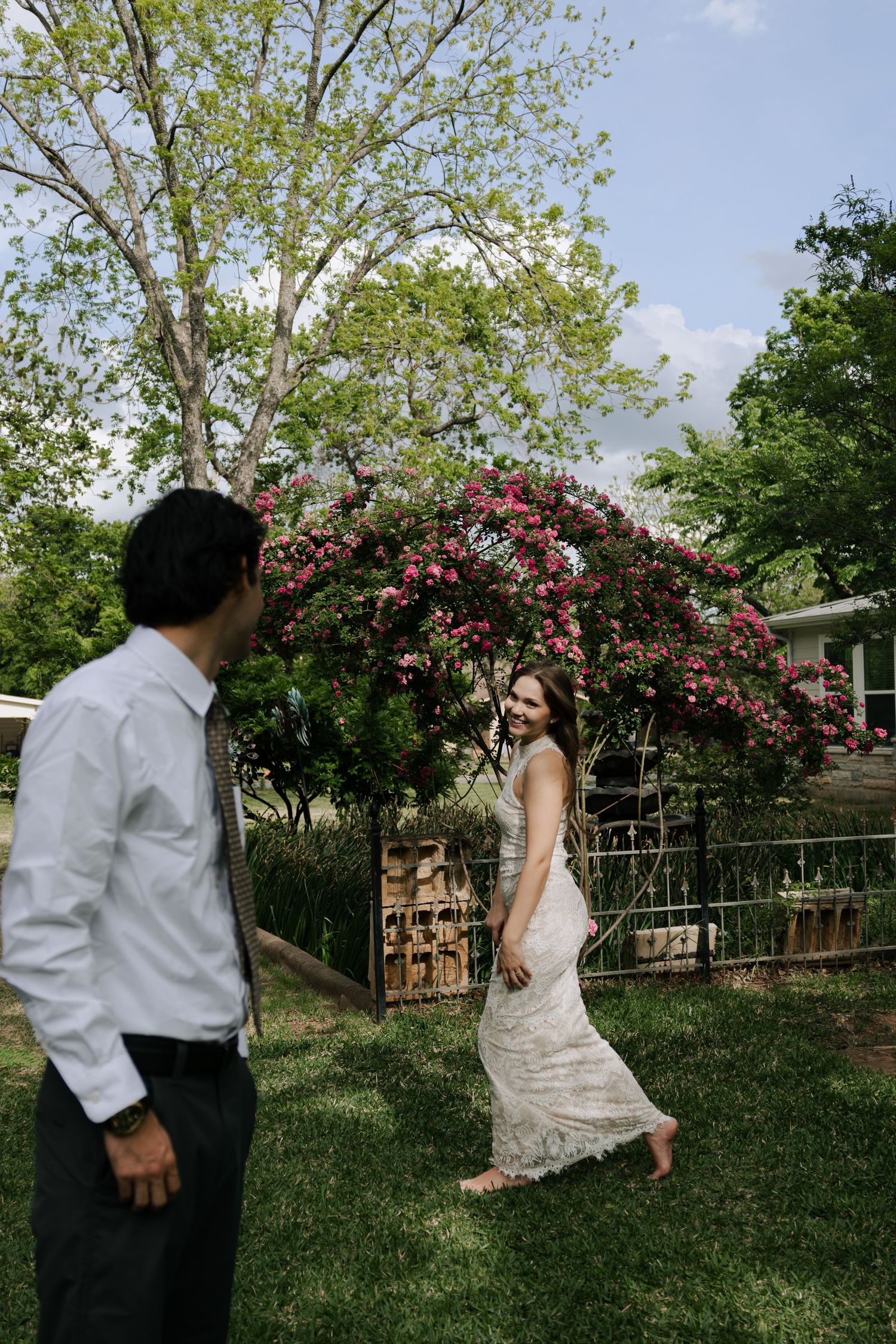Bride in lace wedding dress walking barefoot toward groom in a garden with blooming pink roses.