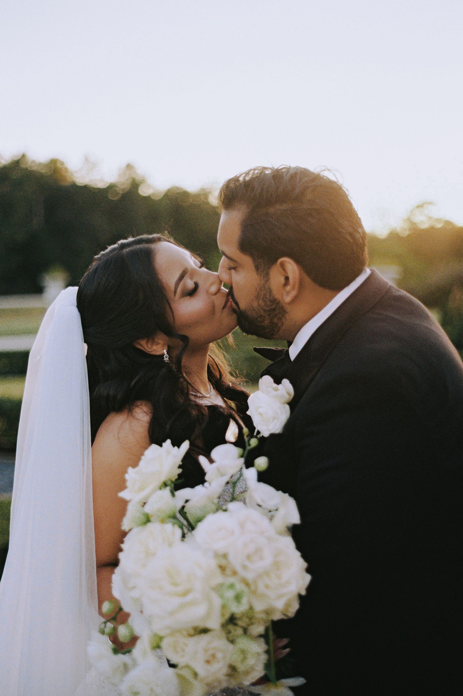 Bride and groom share a romantic kiss at golden hour with a white floral bouquet in foreground.