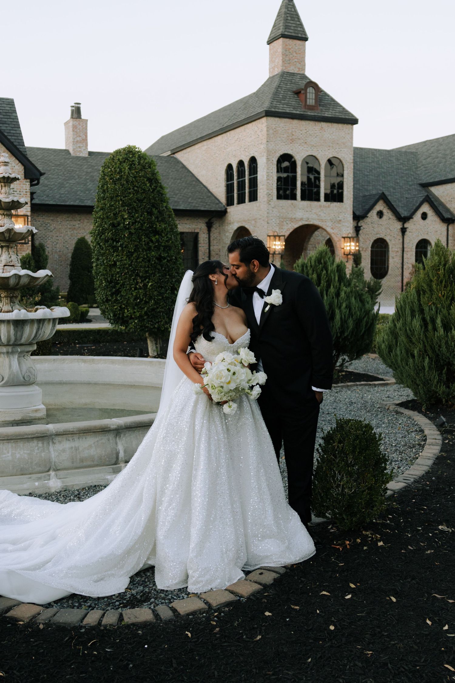 Bride and groom share a kiss near stone fountain outside elegant castle-style wedding venue at dusk.