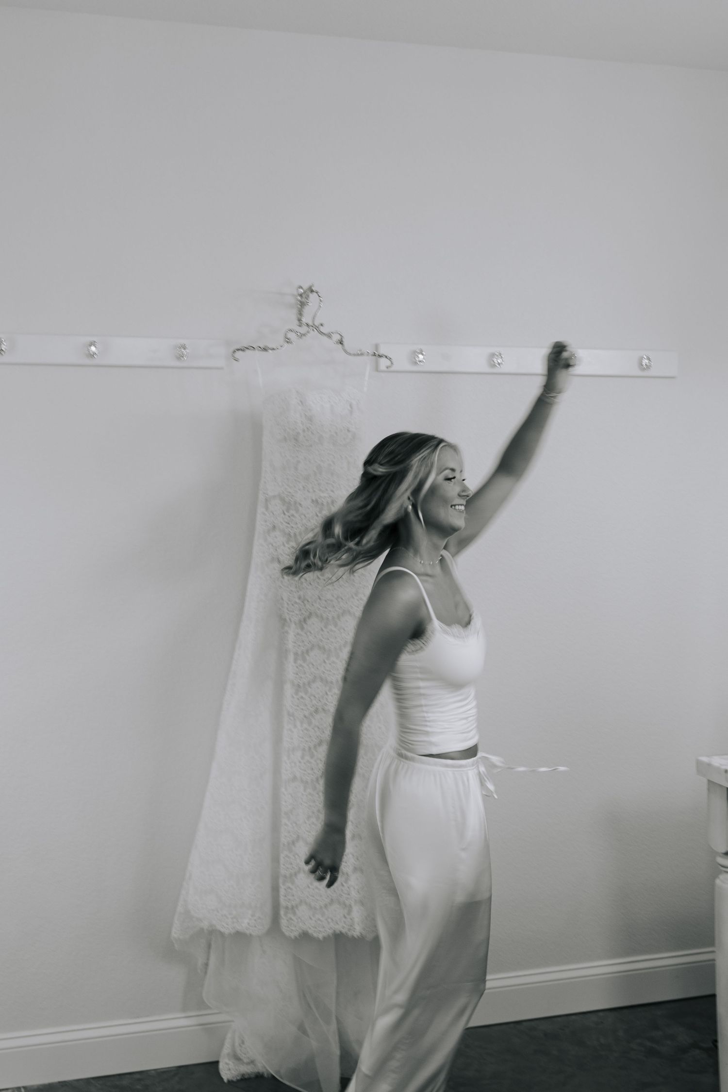 Black and white photo of a smiling bride dancing near her lace wedding dress hanging on the wall.
