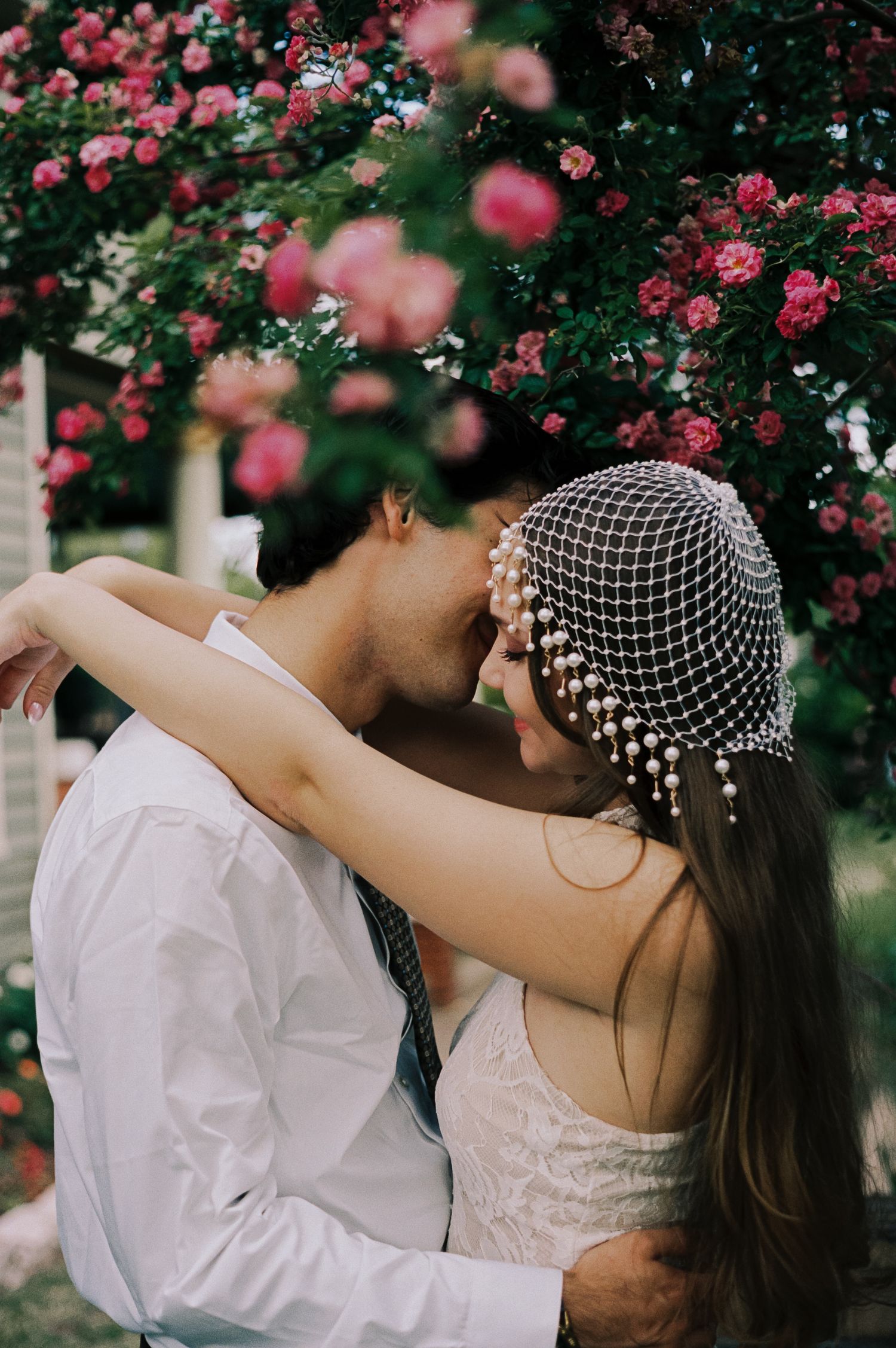 Groom kissing bride wearing a pearl birdcage veil in front of a lush pink rose garden backdrop.