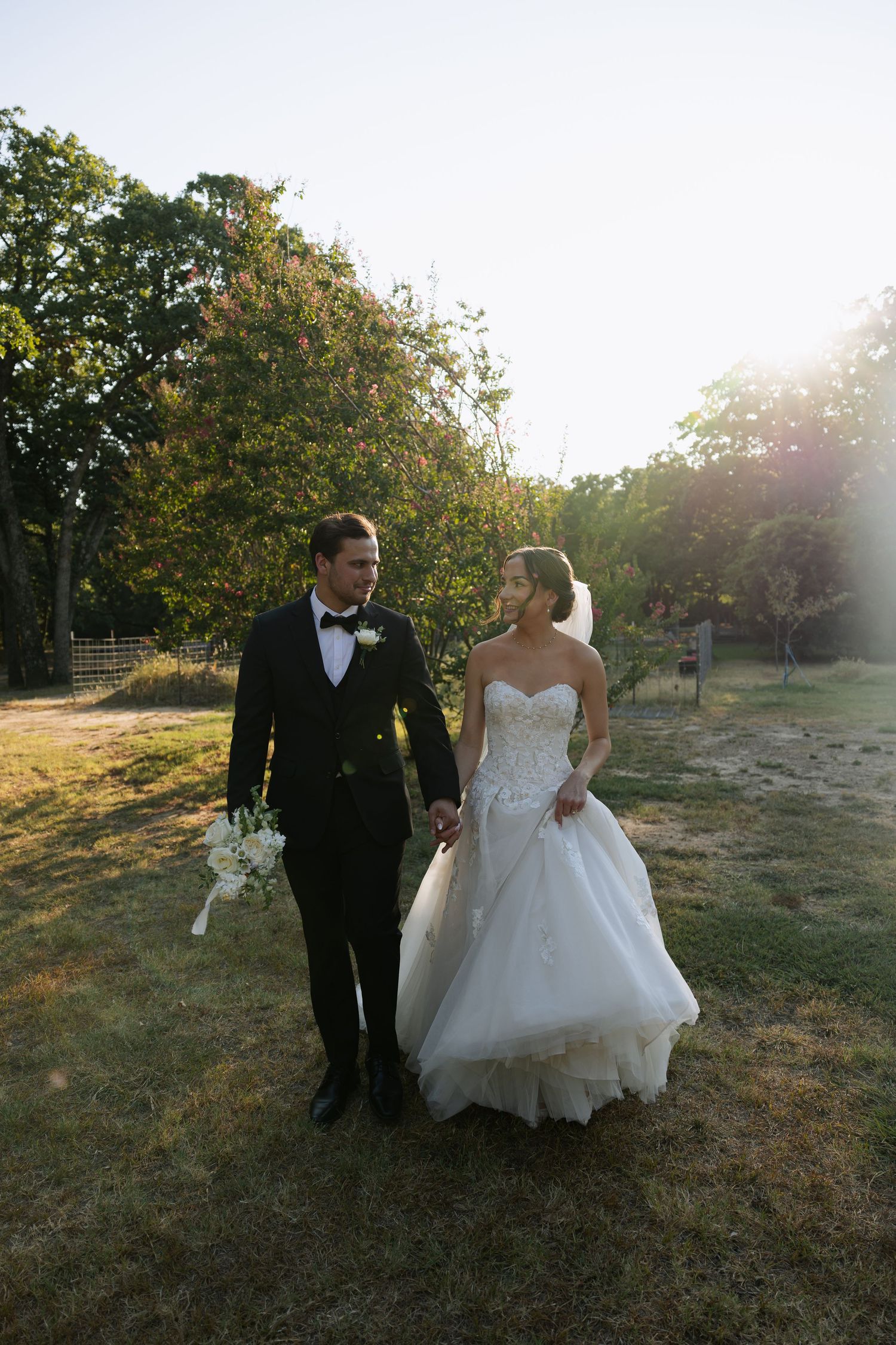 Bride and groom walk hand-in-hand through sunlit garden, she in strapless ball gown, he in black tuxedo.