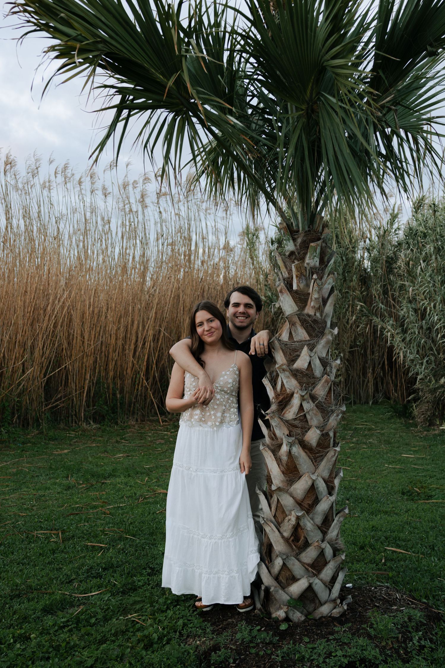 Smiling couple posing together beside a tall palm tree with golden reeds in the background.