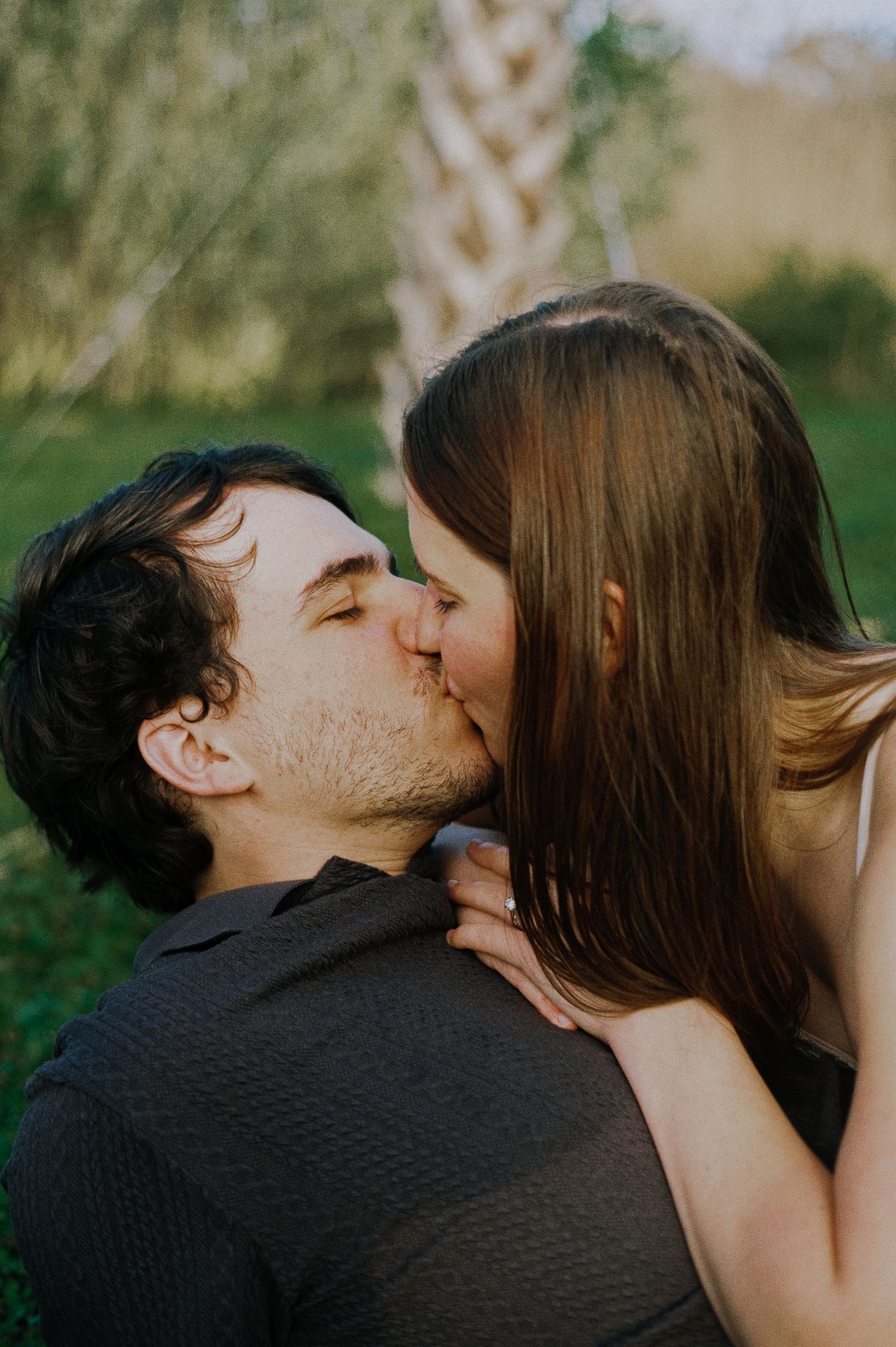 Romantic couple sharing a kiss outdoors with lush green trees in the background.
