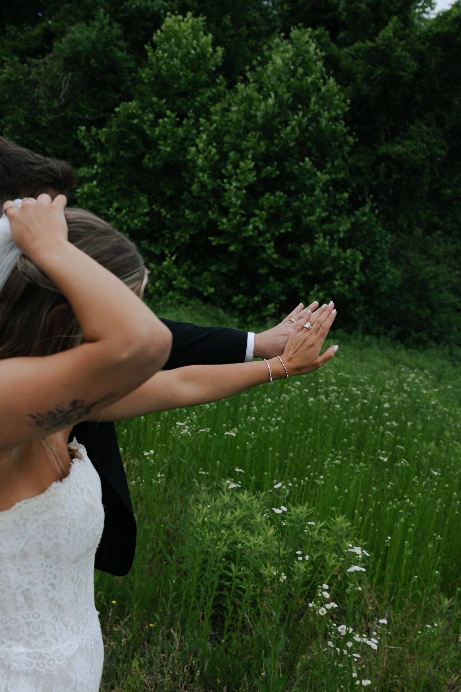 Bride in white lace dress embracing groom outdoors in a lush green meadow with wildflowers.