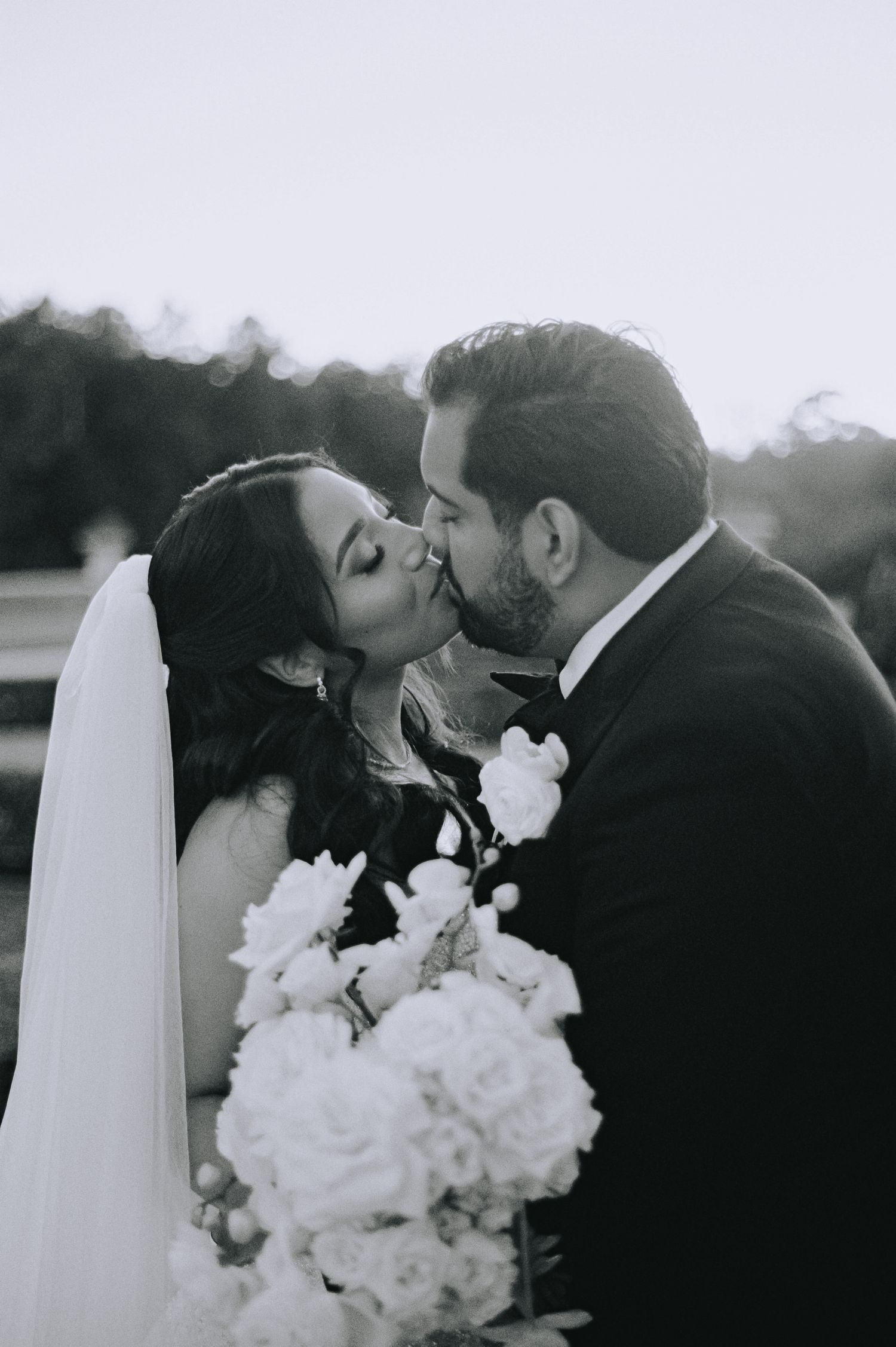 Black and white photo of a bride and groom sharing a kiss, bride holding a white floral bouquet on their wedding day.