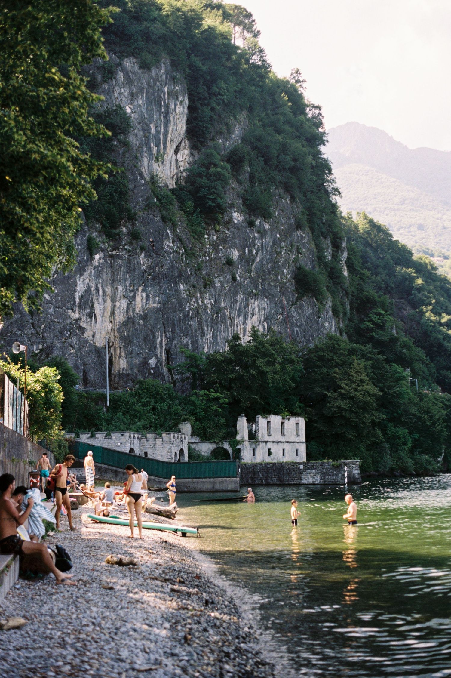 People swimming and relaxing on a rocky shoreline beside a calm lake with a historic building and dramatic cliffs behind.