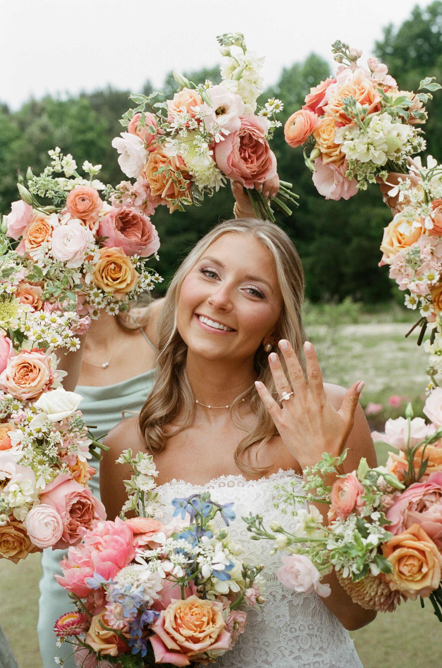 Smiling bride shows off engagement ring framed by large floral arrangement of pink and orange roses.