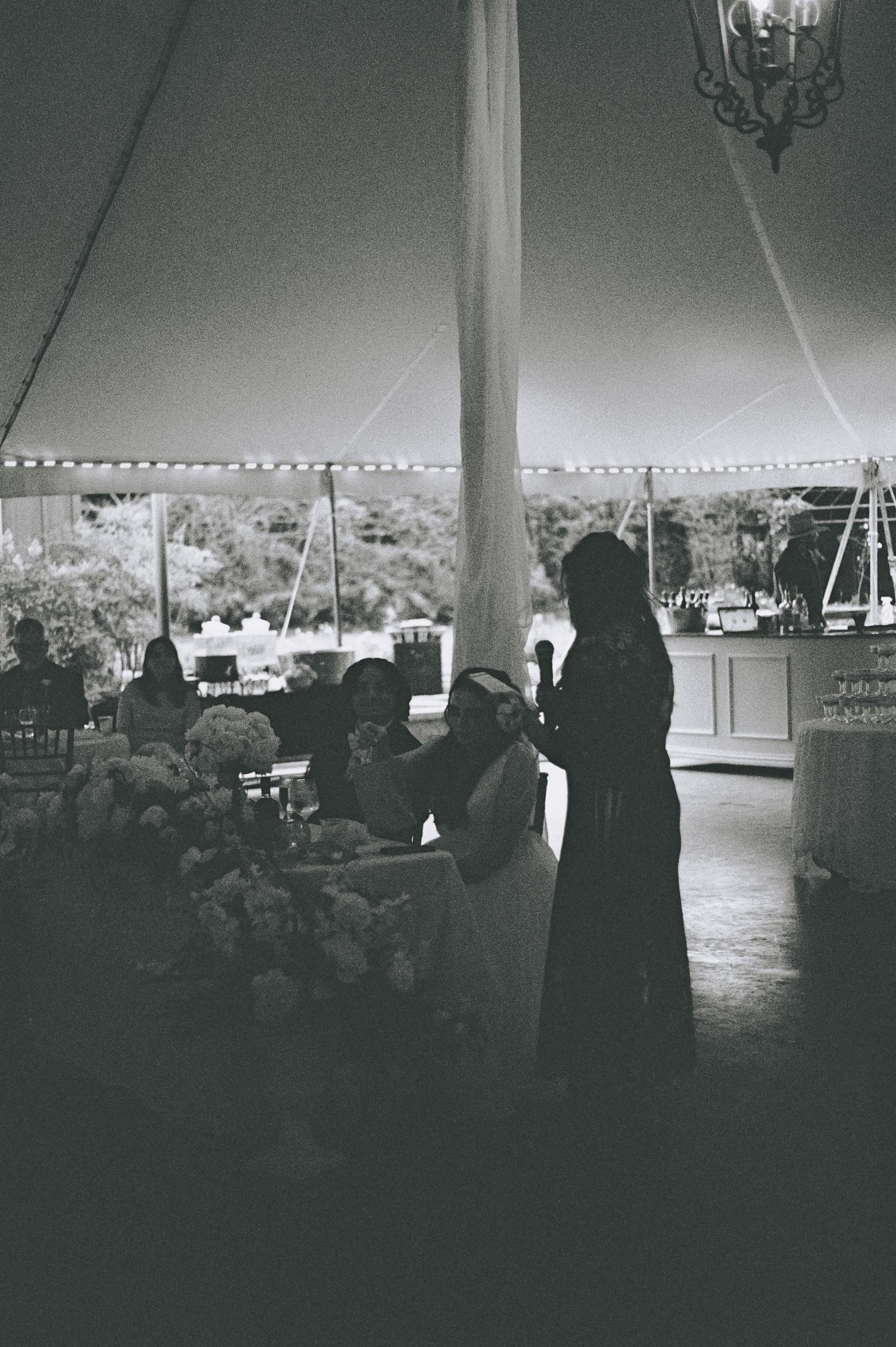 Black and white photo of wedding guests mingling under a elegant tent with string lights at a reception.