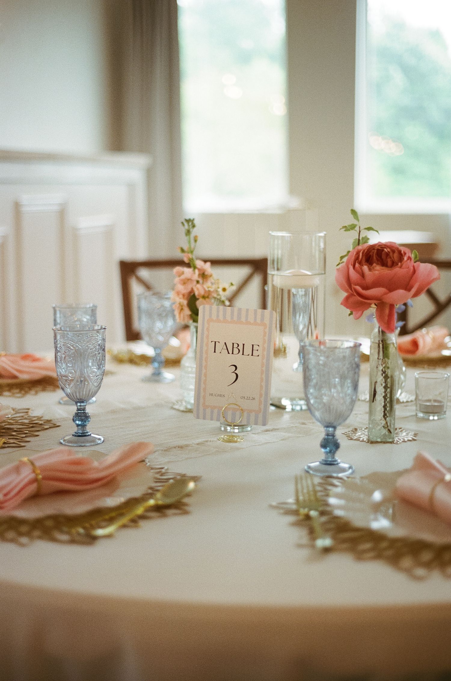 Elegant wedding table setting with blue goblets, pink napkins, coral flowers, and a Table 3 card on a white tablecloth.