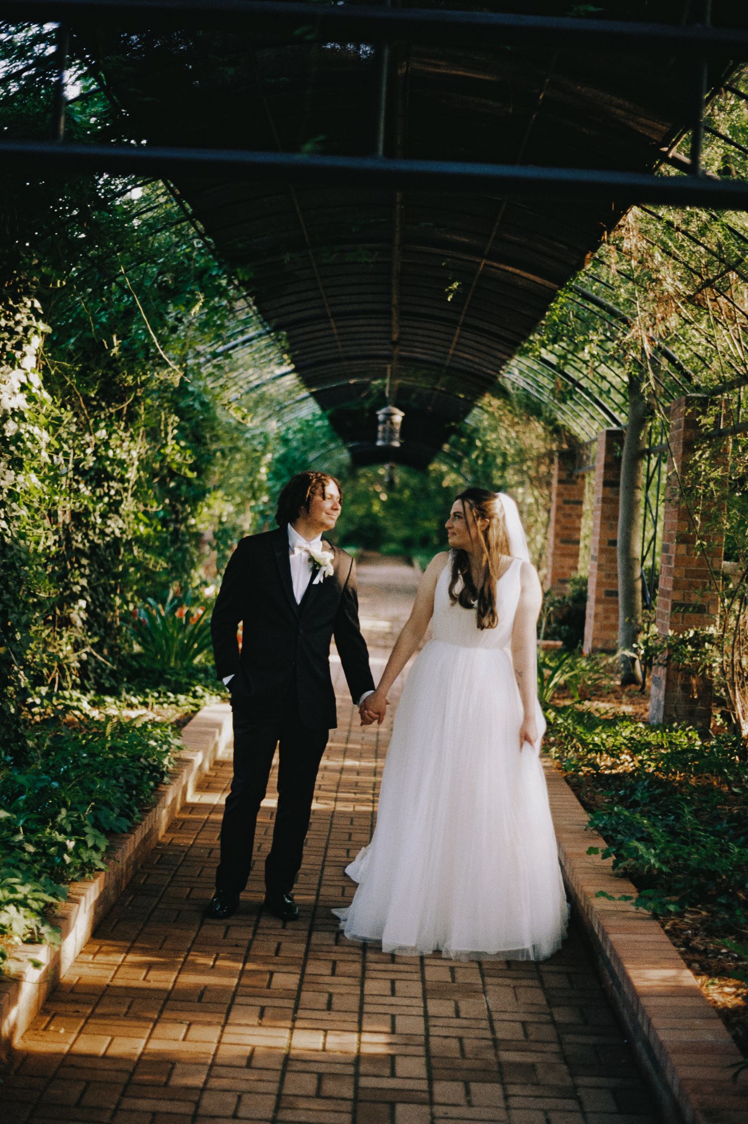 Bride and groom hold hands walking through a lush garden pergola, bride in white gown, groom in black suit.