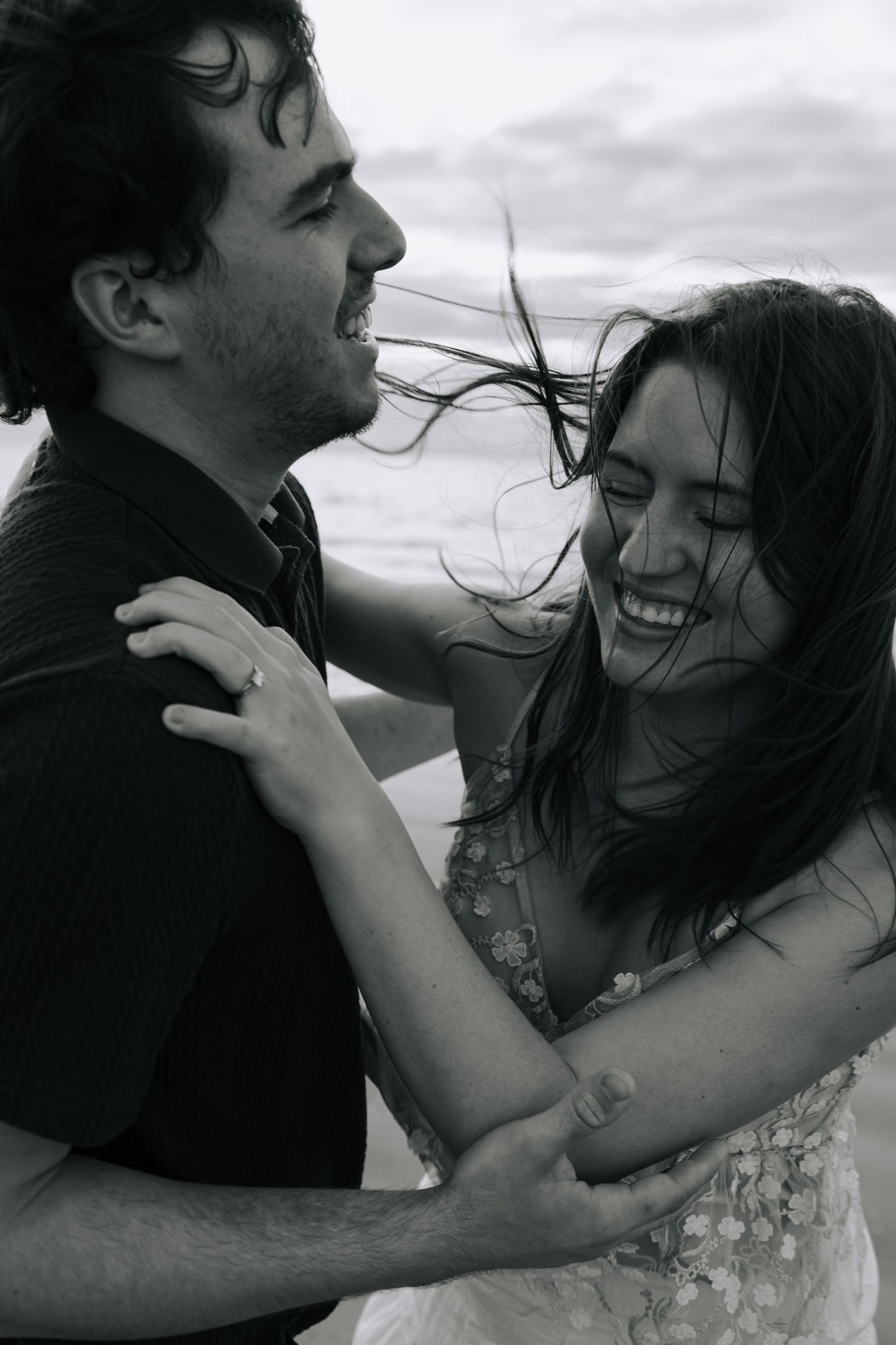 Black and white photo of a joyful couple embracing on a windy beach, woman laughing in a floral dress.