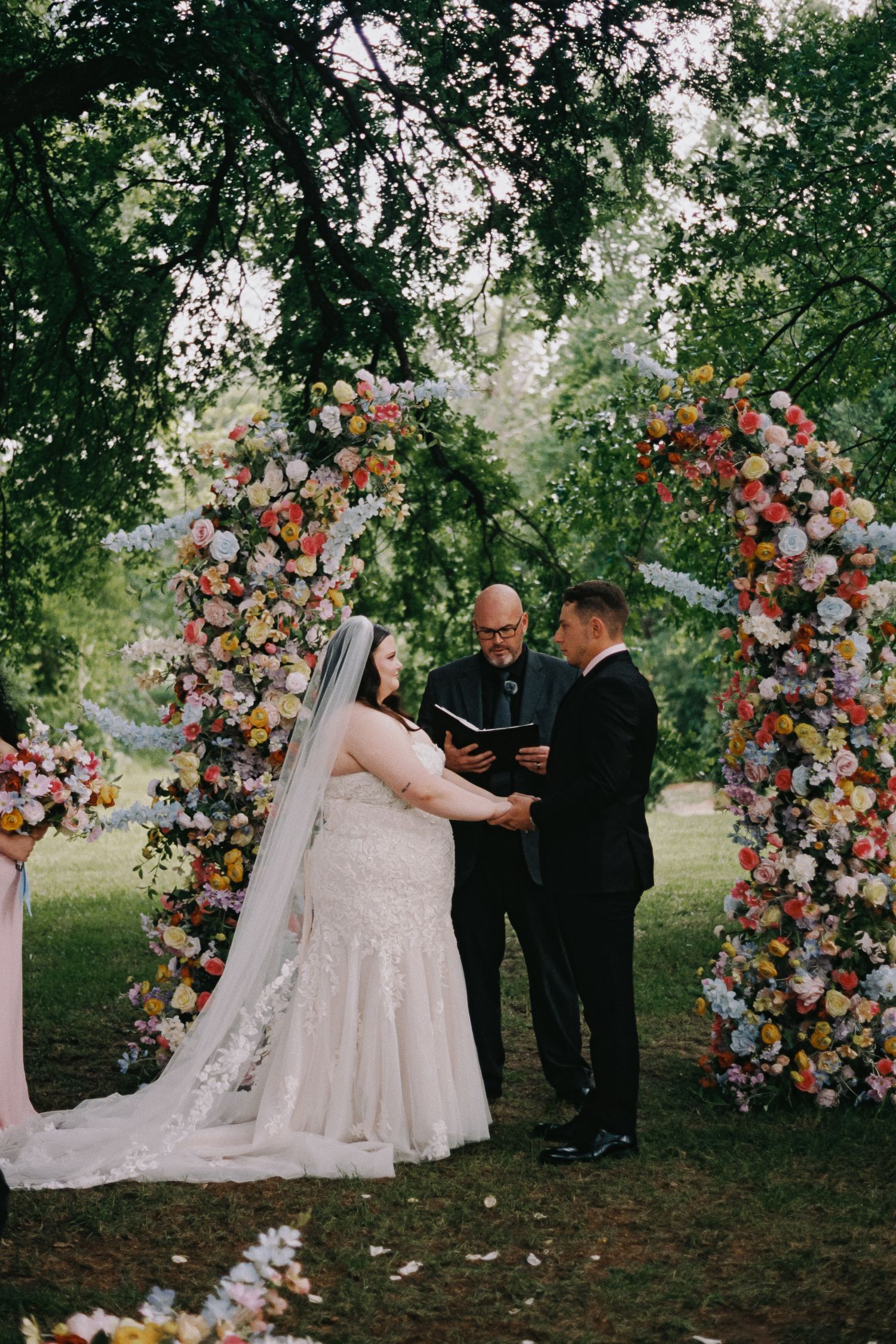 Outdoor wedding ceremony with bride and groom exchanging vows under a colorful floral arch surrounded by lush greenery.