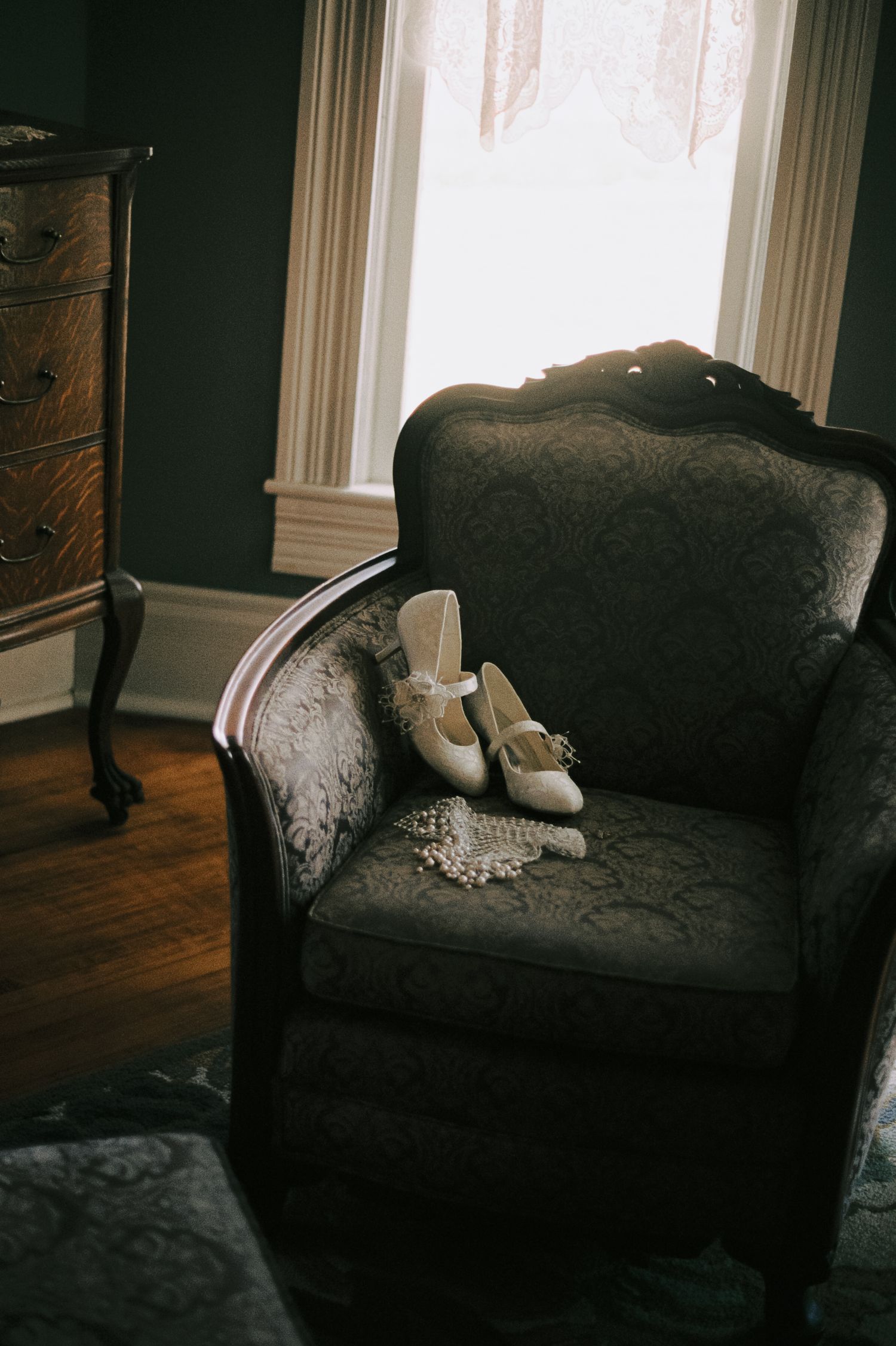 White bridal heels with bow details resting on a vintage tufted armchair near a window.