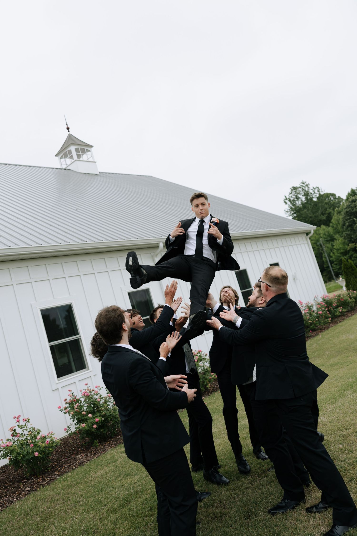Groomsmen tossing groom in the air outside a white barn wedding venue.