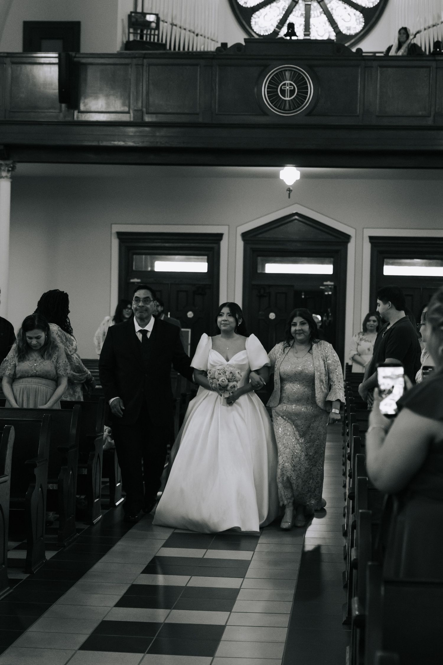 Black and white photo of a bride walking down the church aisle with family during her wedding ceremony.
