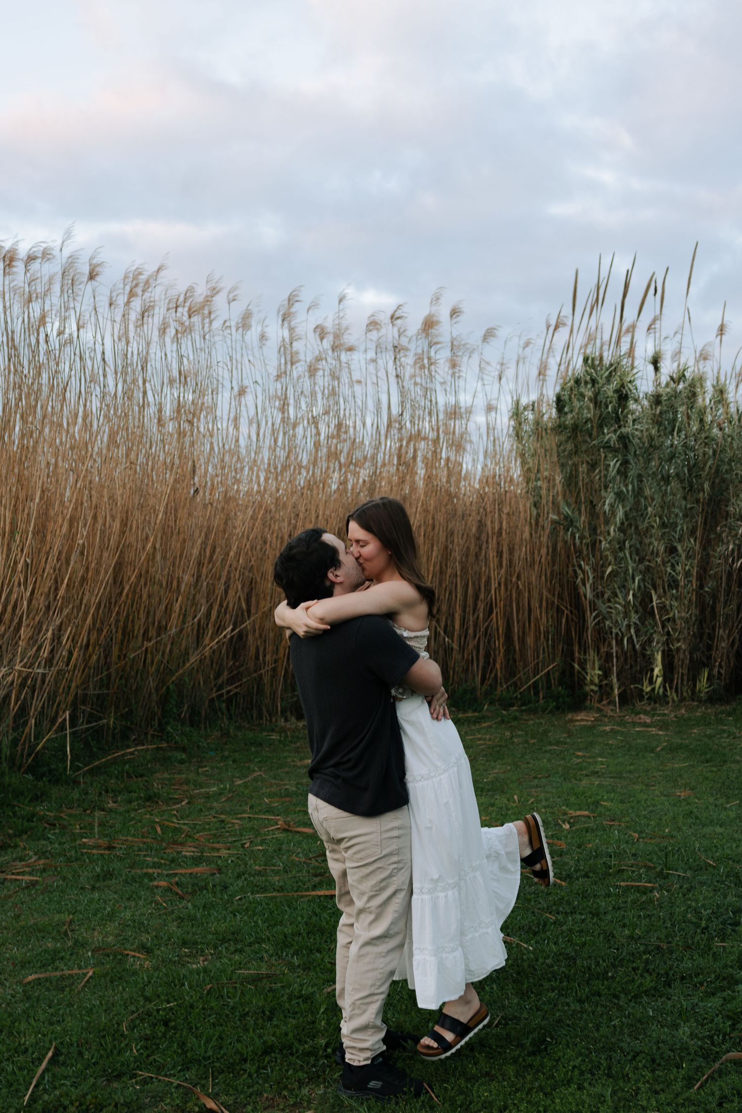 Groom lifting and kissing bride in white dress in front of tall golden reeds at sunset engagement shoot.