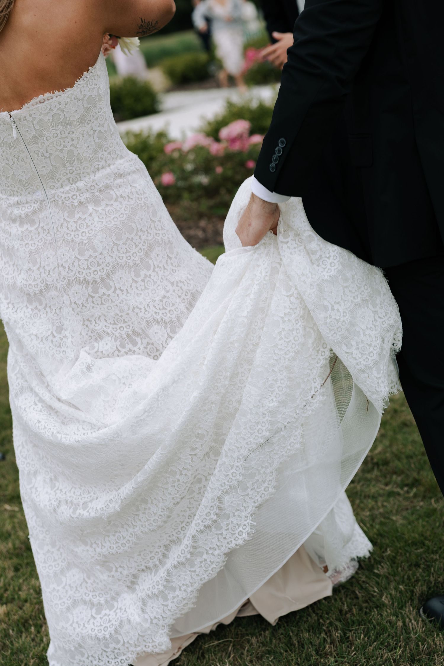Groom in black suit lifting the train of a white lace wedding dress outdoors near a garden.