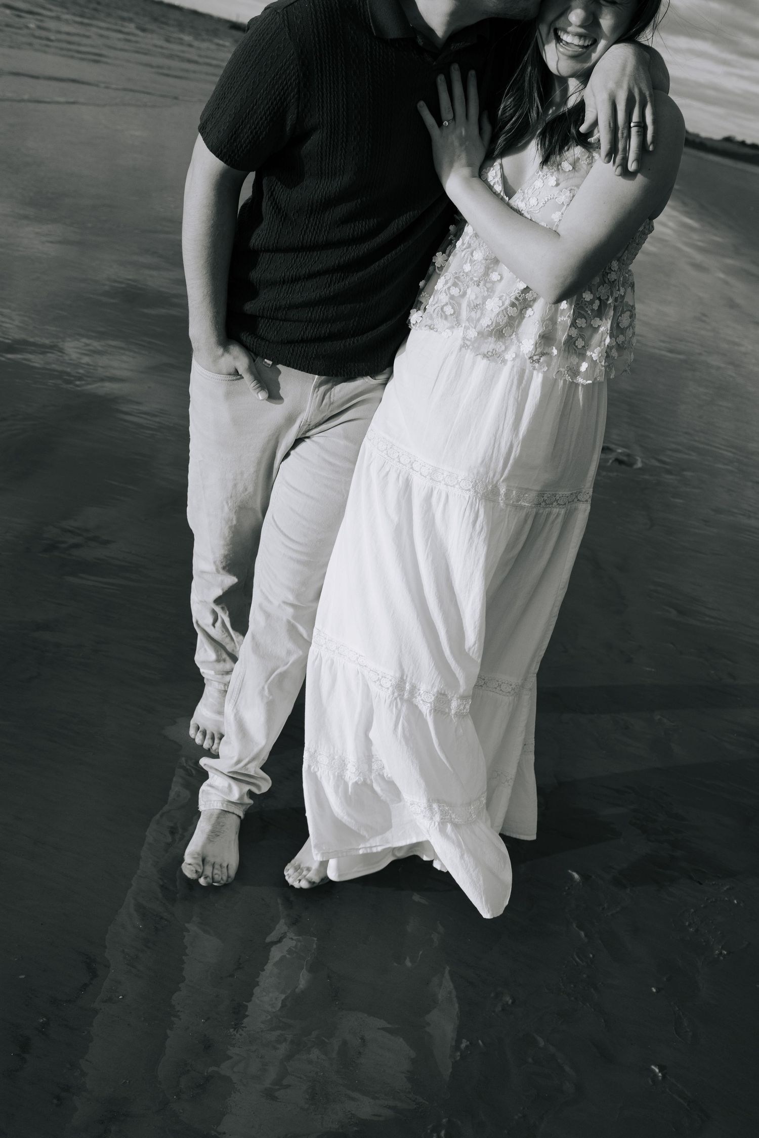 Black and white photo of couple standing together on a reflective beach, woman in flowing white dress.
