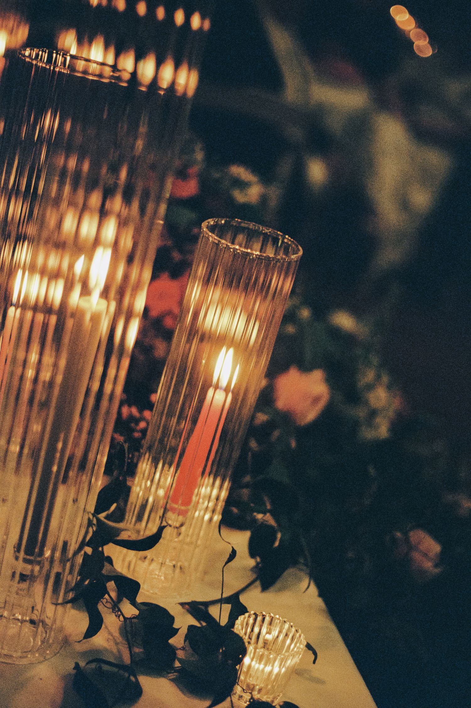 Elegant tall ribbed glass candle holders with glowing candles and floral greenery on a reception table.