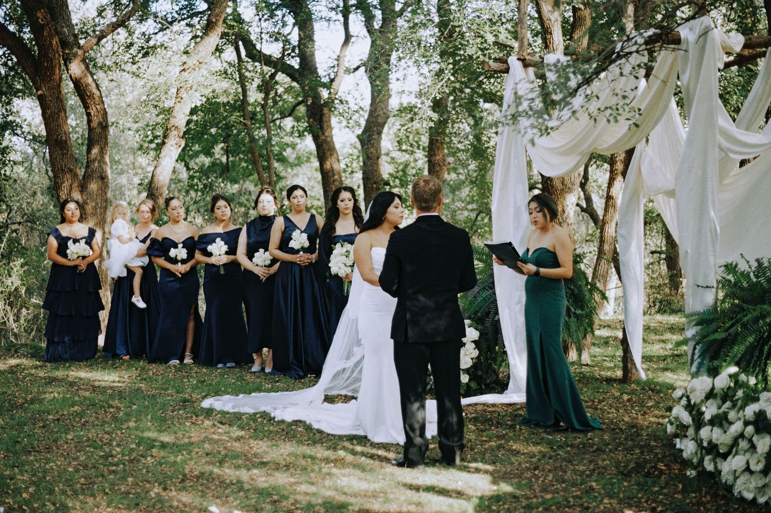 Outdoor wedding ceremony with bridesmaids in navy dresses standing beneath draped white fabric and trees.