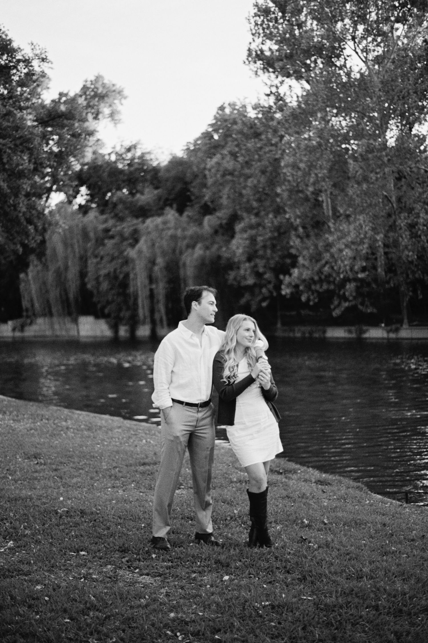 Black and white photo of a couple walking hand in hand by a serene lake surrounded by lush trees.