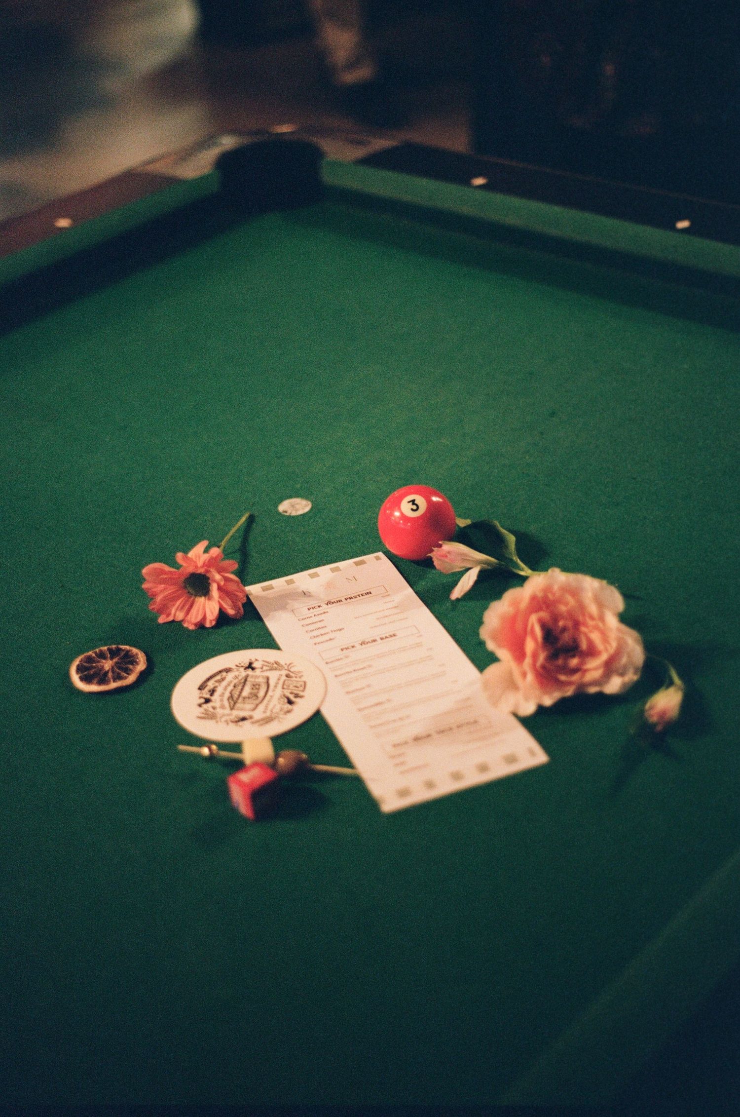Pool table with flowers, a red billiard ball, dried orange slice, and a menu card arranged decoratively on green felt.