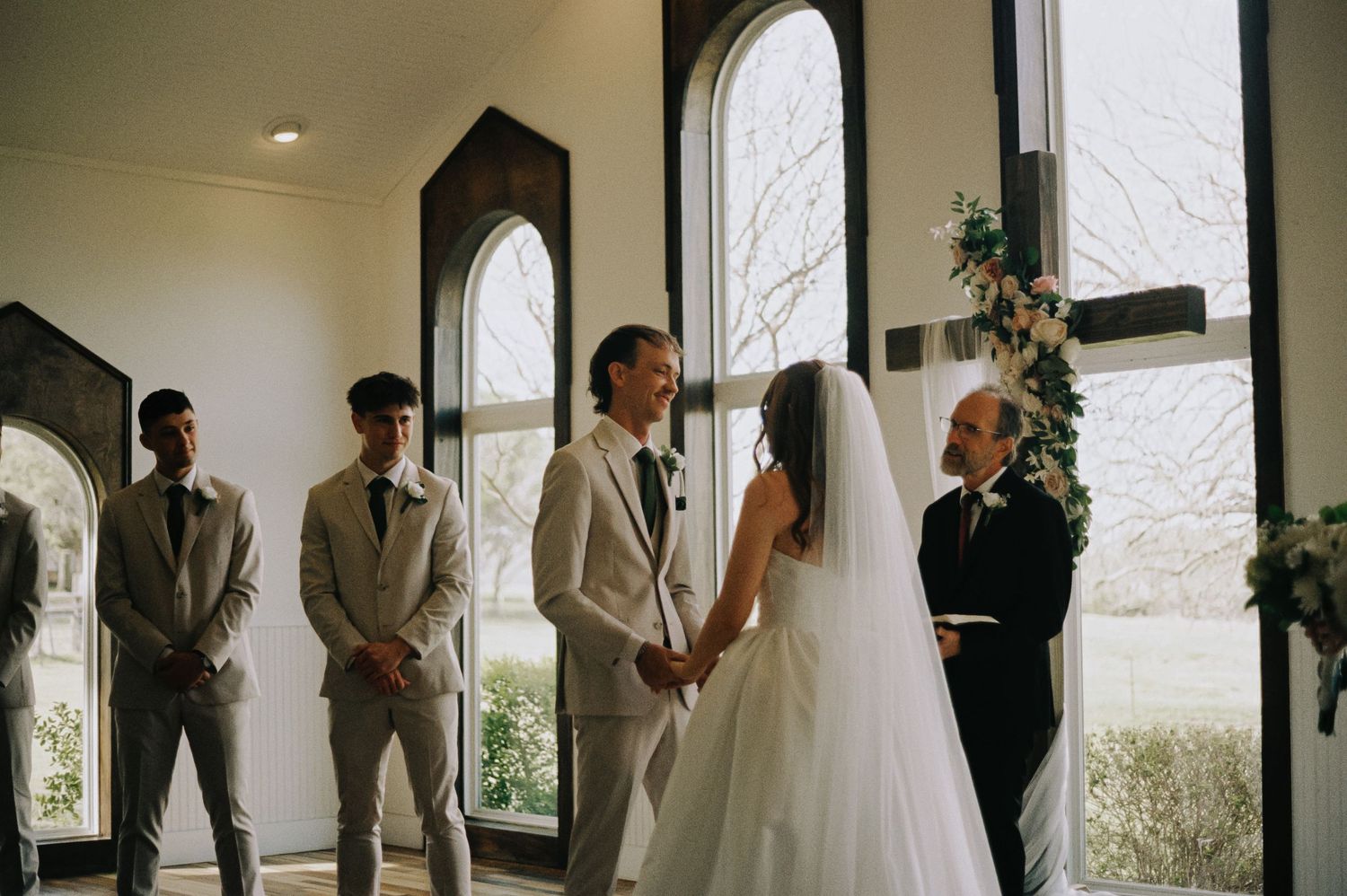 Bride and groom holding hands at wedding altar with groomsmen and officiant in elegant chapel.