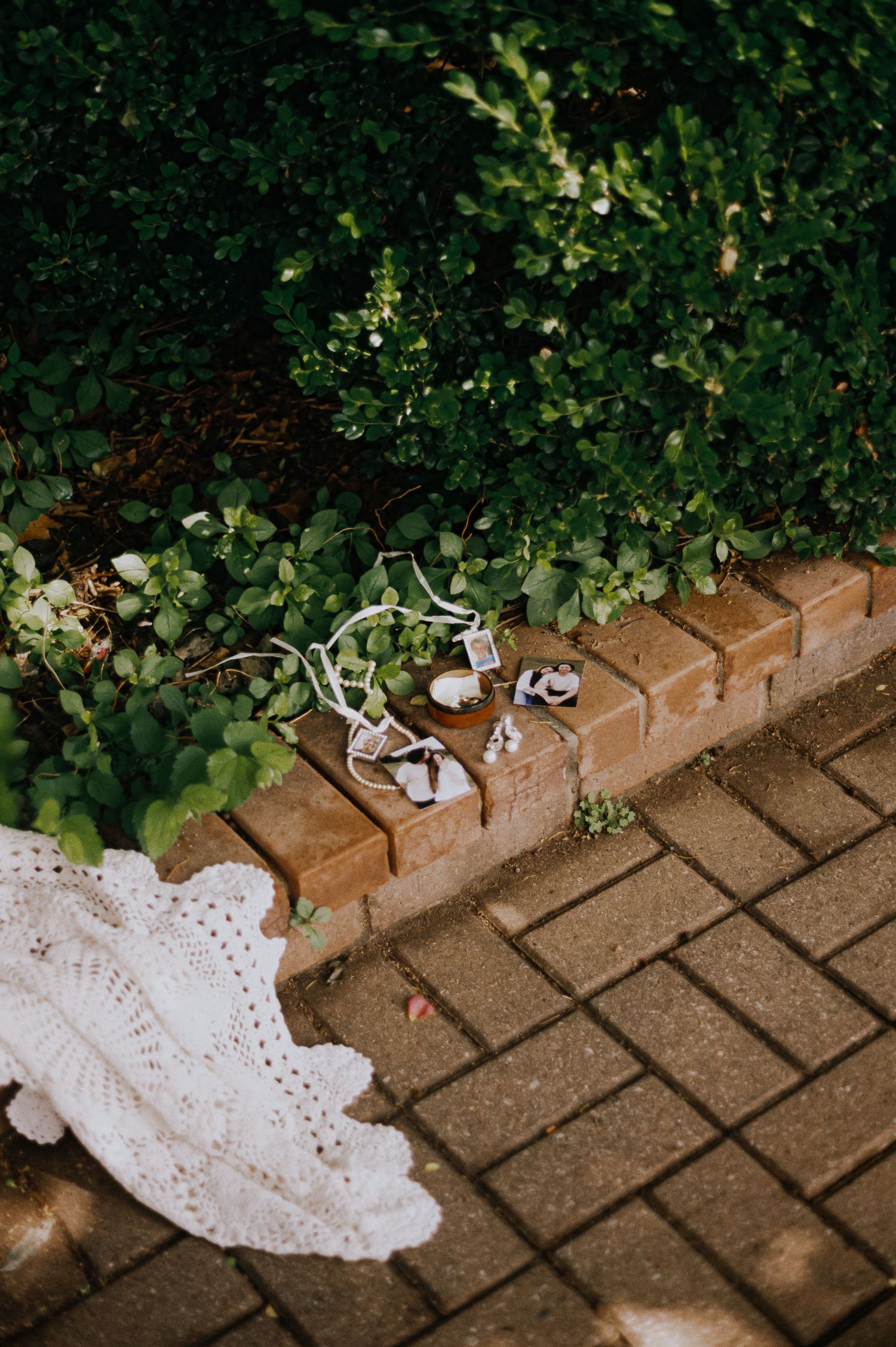 Wedding accessories and white lace dress laid on brick garden edging surrounded by lush green foliage.