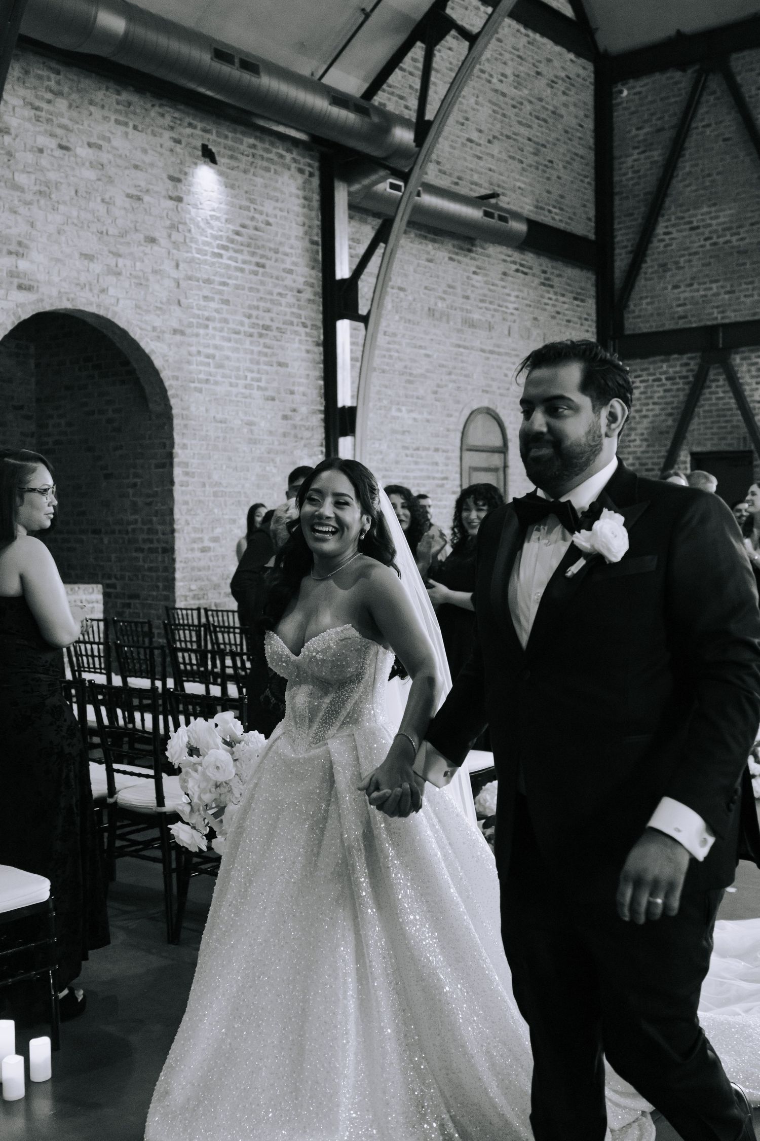 Black and white photo of a joyful bride and groom walking down the aisle after their wedding ceremony in a brick venue.