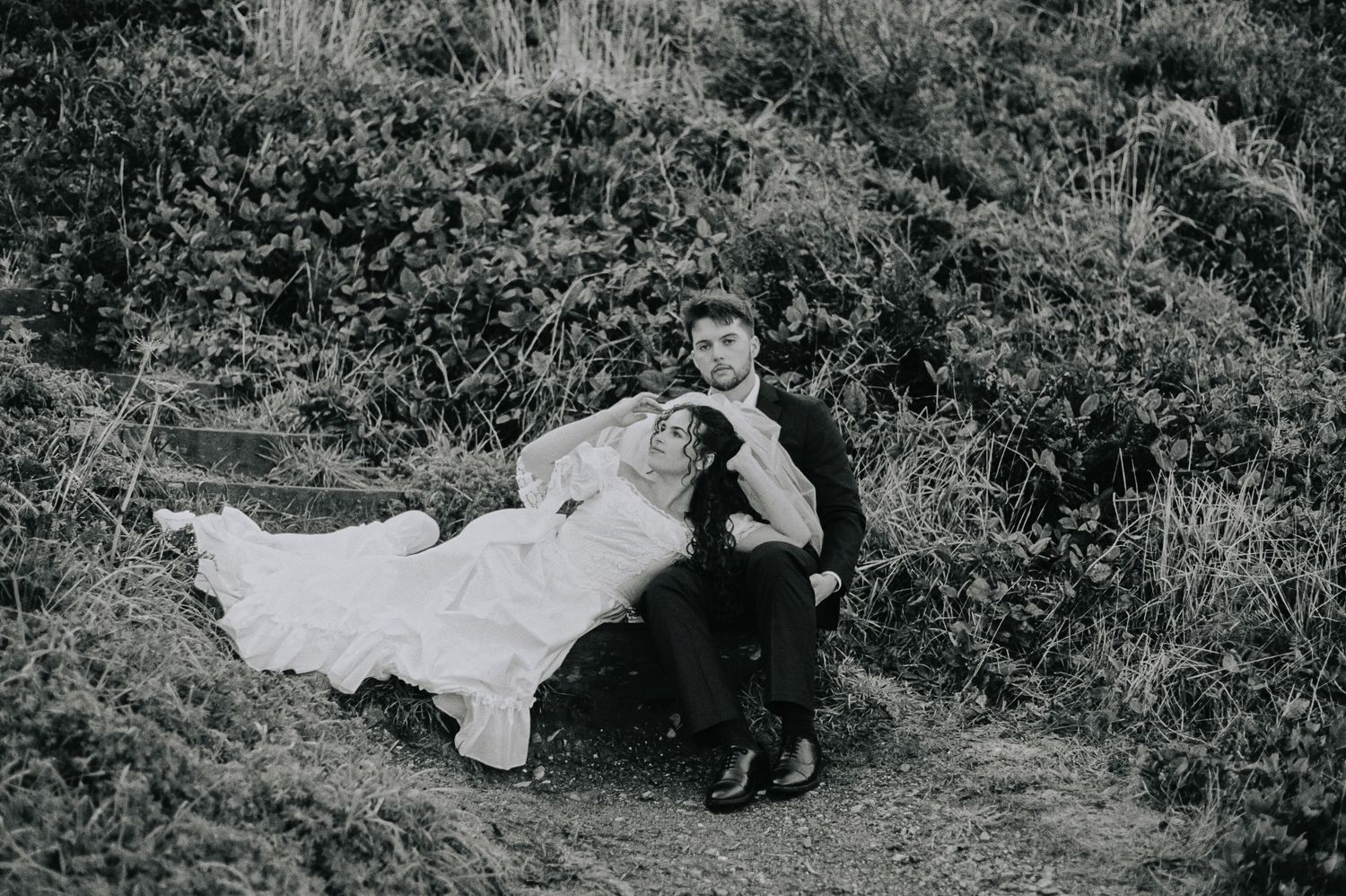 Black and white photo of bride and groom sitting together on a grassy hillside in wedding attire.