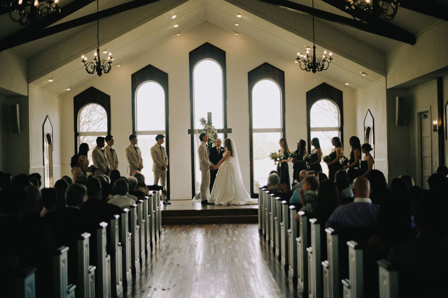 A couple exchanges vows at a chapel wedding ceremony with white pews, arched windows, and chandeliers.