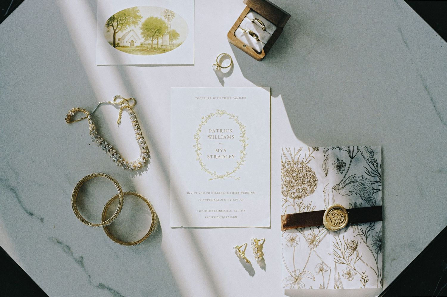 Elegant wedding flat lay with gold rings, pearl bracelet, botanical stationery suite, and wax seal on marble surface.