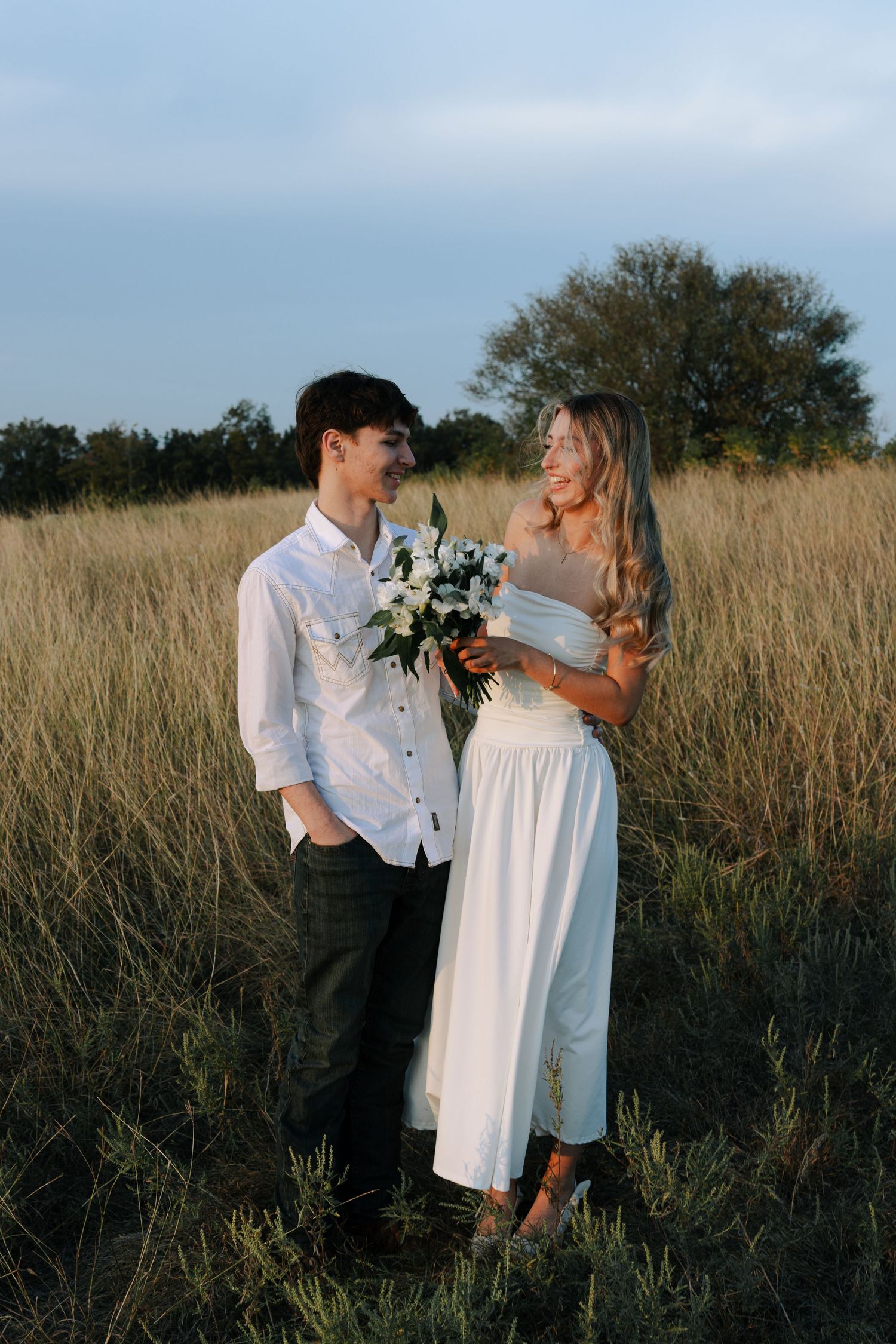 Young couple standing in a golden field at sunset, bride holding white bouquet in white dress.