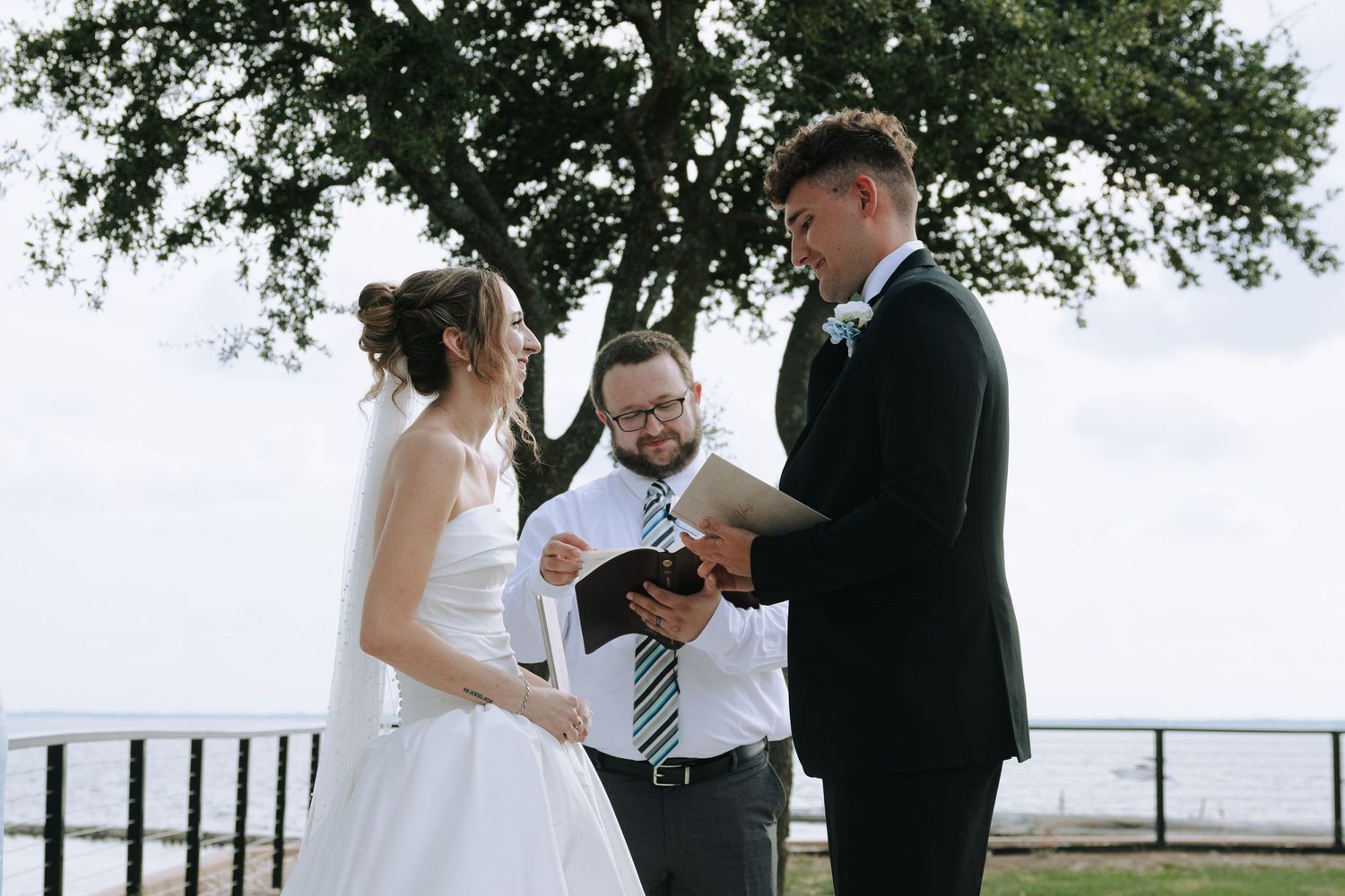 Outdoor wedding ceremony with bride and groom exchanging vows with officiant near waterfront tree.