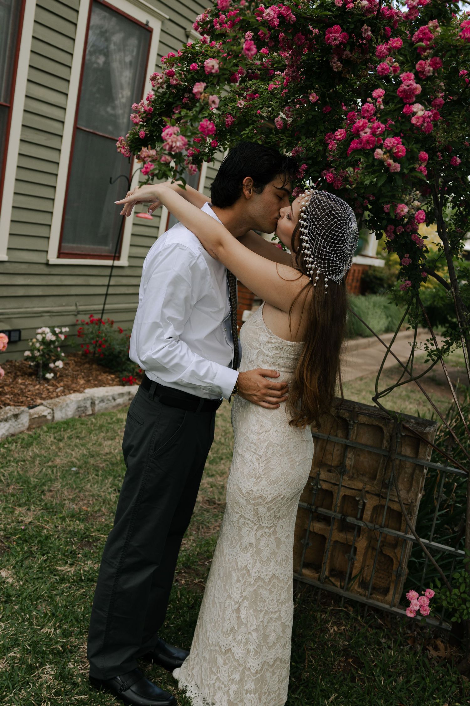 Bride and groom share first kiss under pink rose bush arch beside house.