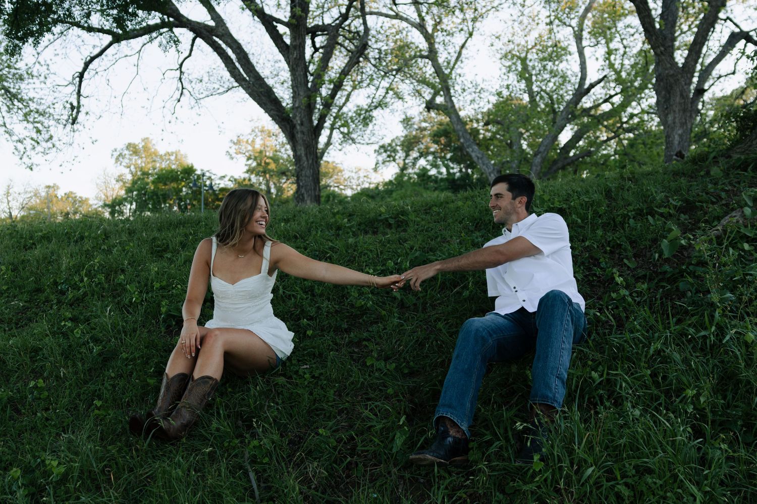 Couple holding hands on a grassy hillside, woman in white dress and boots, man in white shirt and jeans.