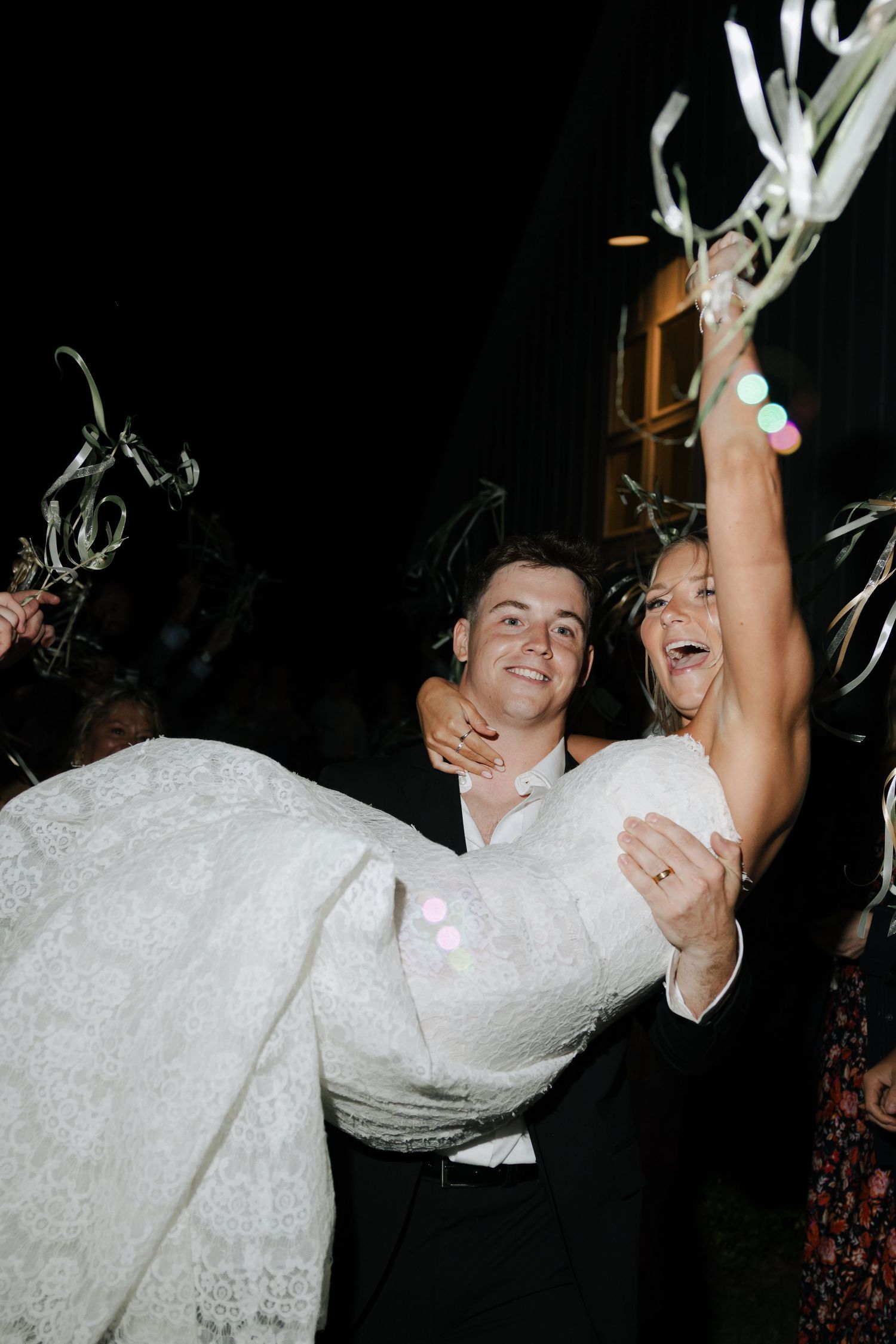 Groom carrying bride in lace wedding dress during festive nighttime wedding send-off with ribbon wands.
