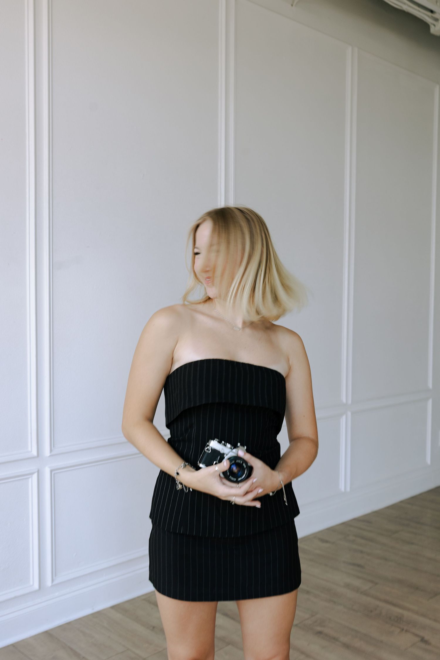 Blonde woman in black strapless mini dress holding camera against white paneled wall.