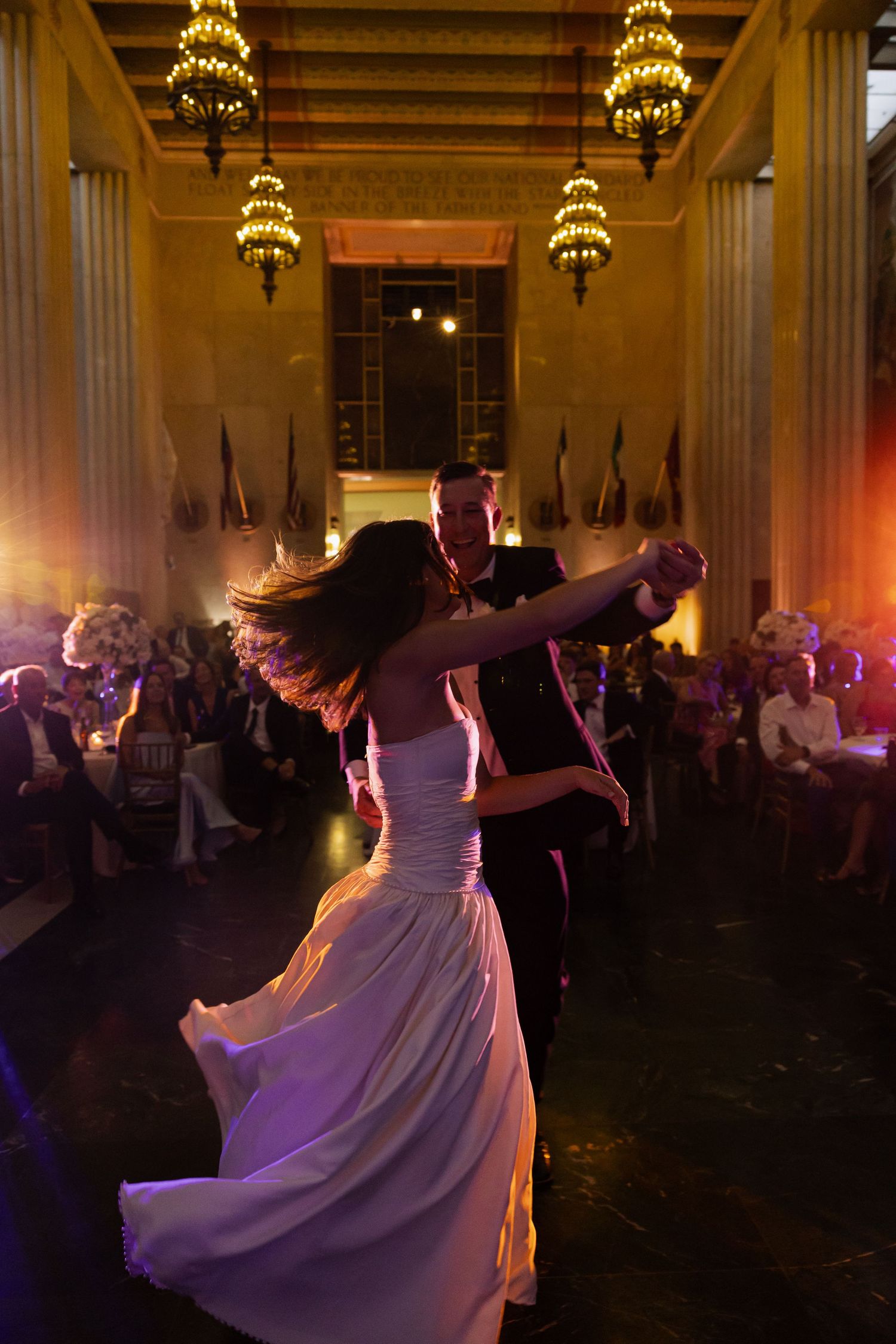 Bride and groom share their first dance in an elegant ballroom with chandeliers and guests seated nearby.