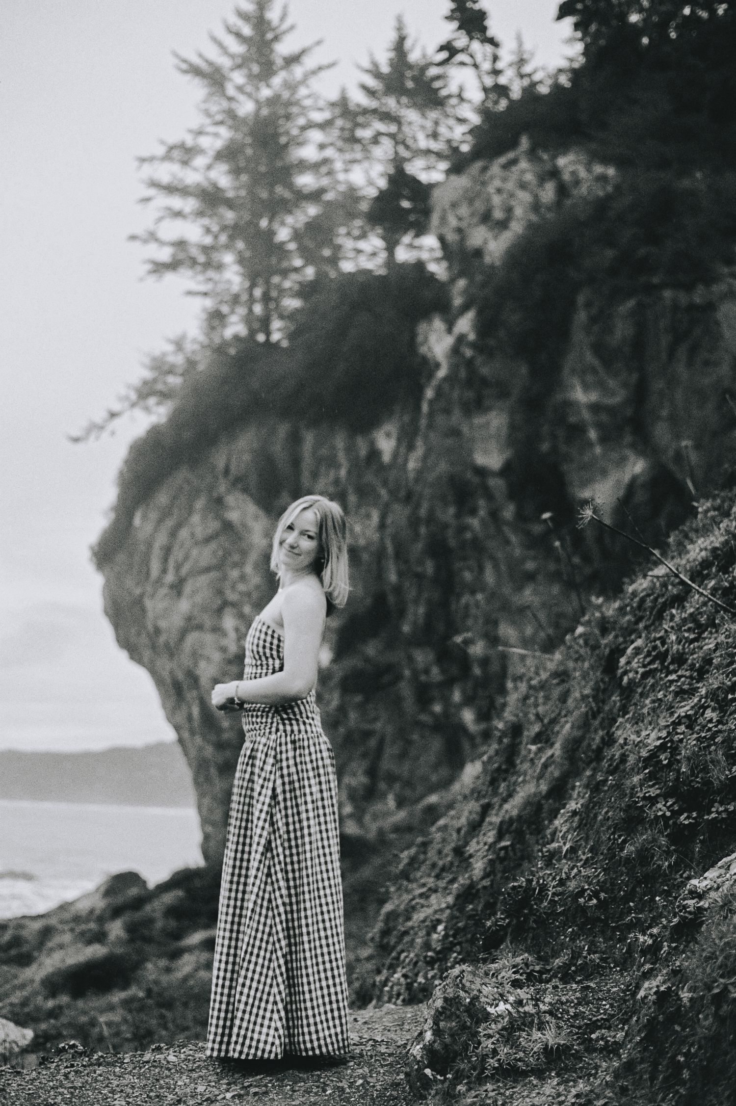 Woman in gingham maxi dress smiling beside rocky coastal cliff with ocean in background.