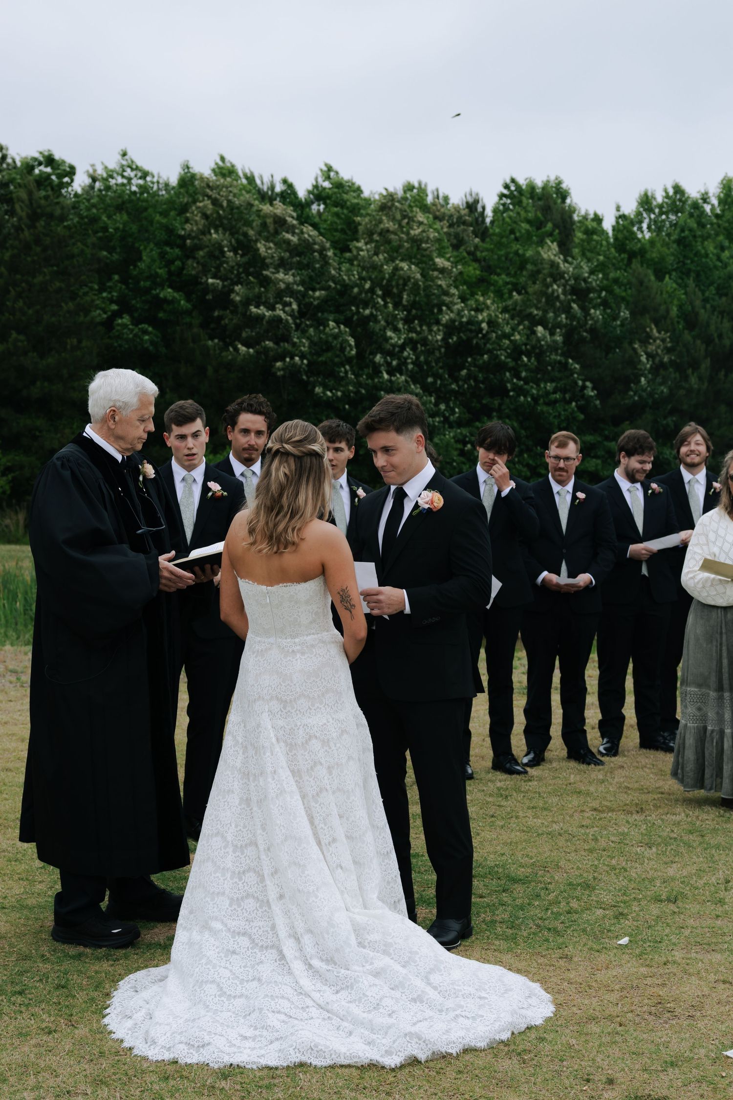 Outdoor wedding ceremony with bride and groom exchanging vows before officiant and groomsmen on lush green lawn.