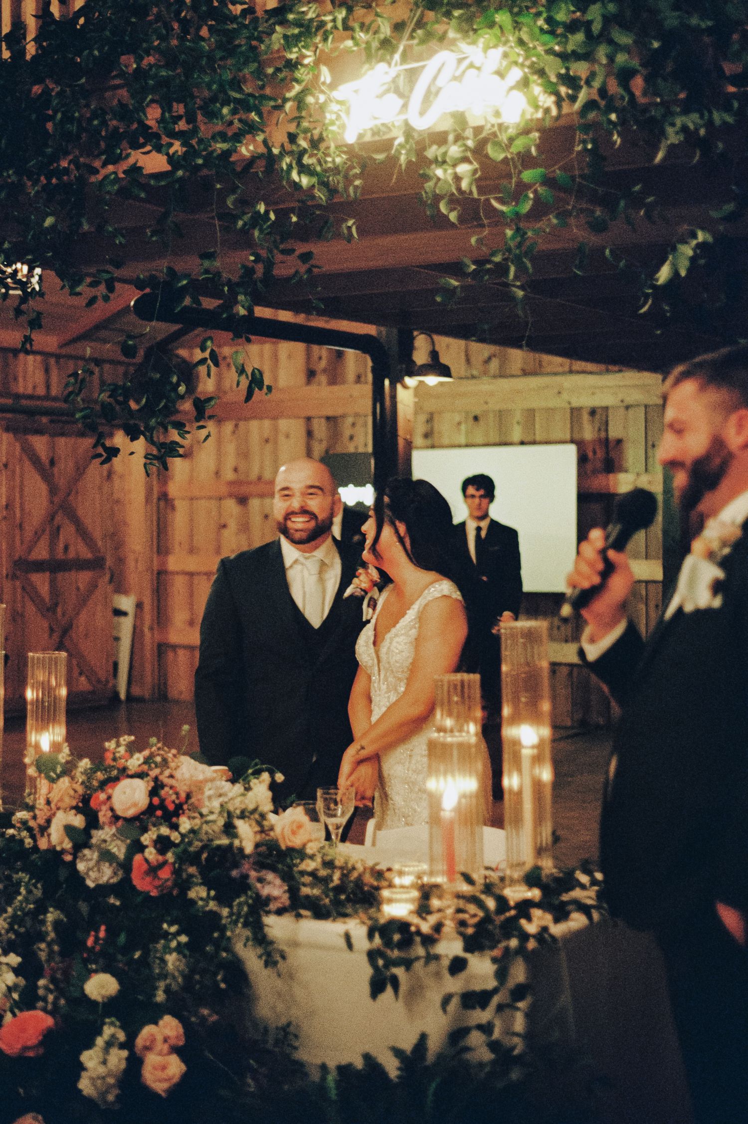 Bride and groom smiling at sweetheart table surrounded by candles and florals in a rustic barn venue.