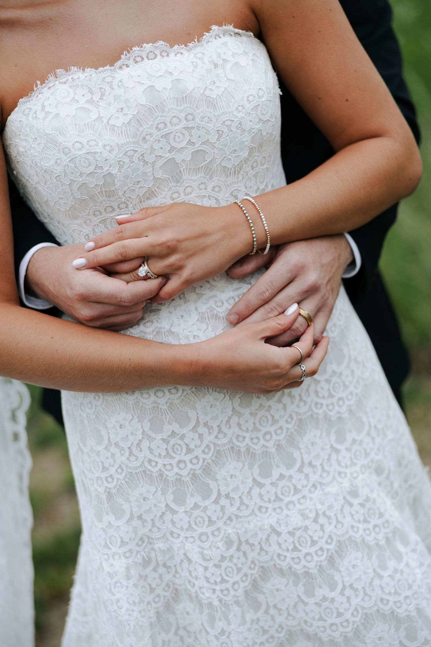 Close-up of bride and groom hands showing wedding rings on white lace strapless wedding gown.